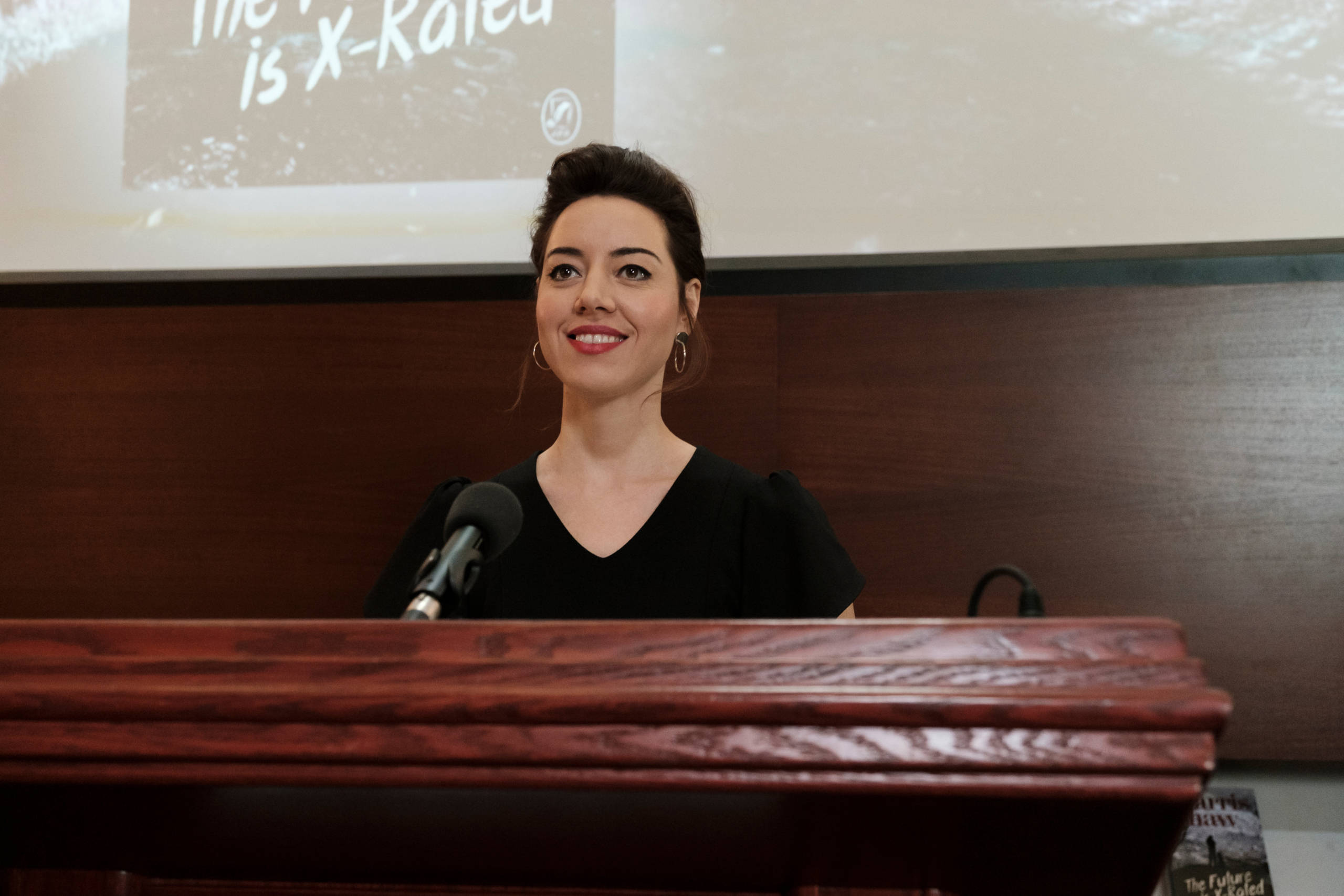 奧布萊·普拉薩飾演露西·斯坦布里奇 壁紙 — 一位身穿黑色上衣的女性站在拋光木製講台後面，手持麥克風，溫暖地微笑。
