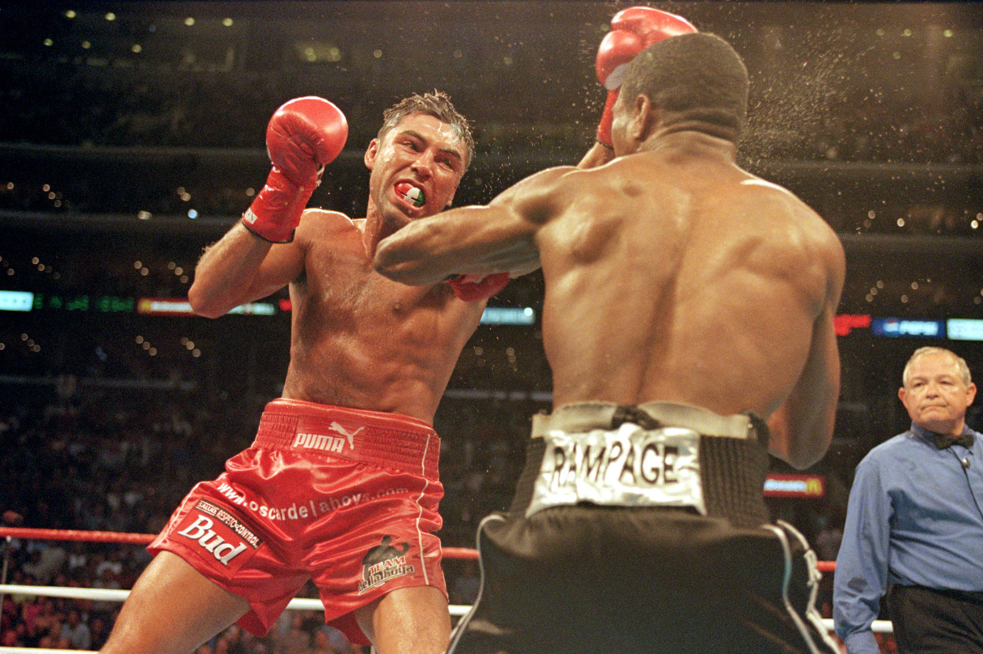 奧斯卡·德·拉·霍亞對決夏恩·莫斯利 壁紙 — 一名穿著紅色短褲的拳擊手在燈光明亮的擂台上，因承受對手的重拳而扭曲表情。