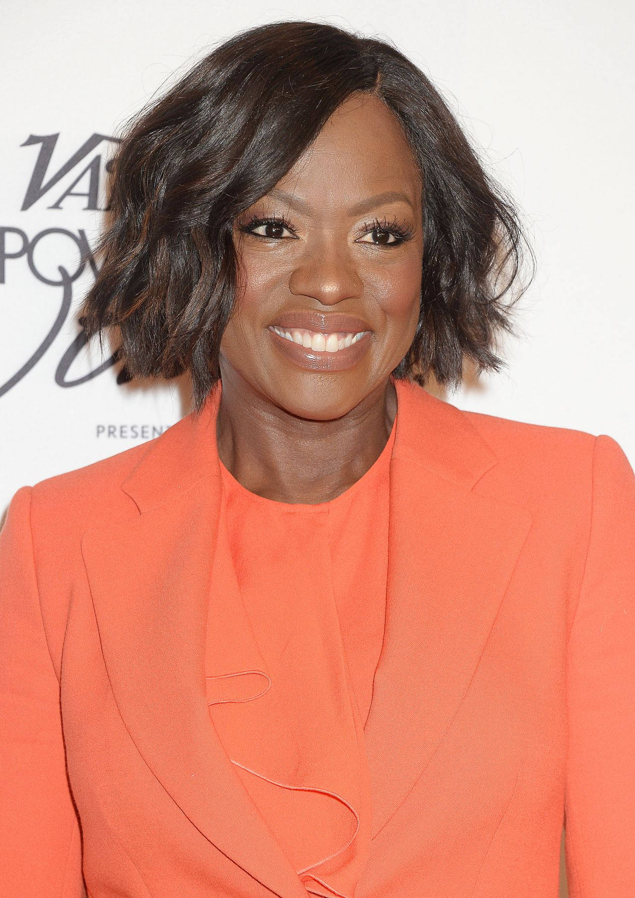 奧斯卡得主維奧拉·戴維斯為《Vanity Fair》拍攝時展現優雅風采 壁紙 — 一位容光煥發、笑容溫暖的女性，在白色活動背景前穿著大膽的橙色西裝外套。