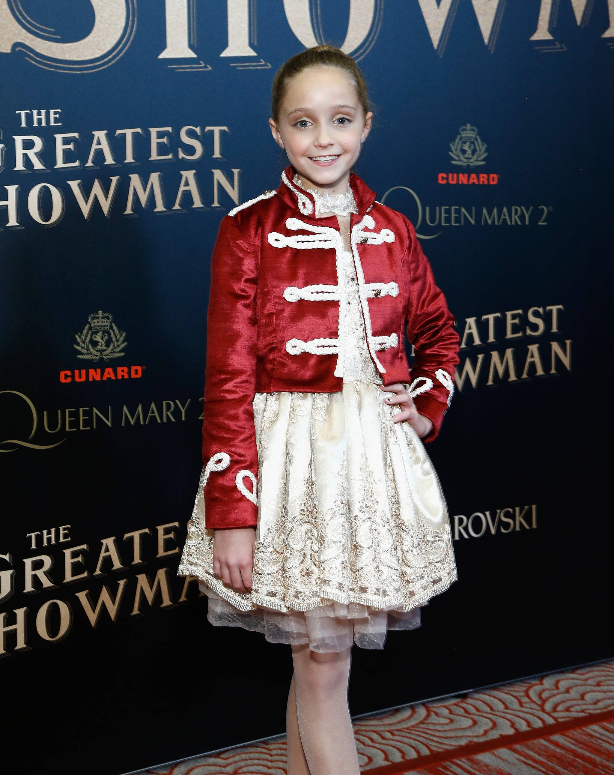 奧斯汀·強森在《 Greatest Showman 》 壁紙 — 一個穿著紅色天鵝絨外套和奶油色蕾絲連衣裙的小女孩在電影首映紅地毯活動中微笑。