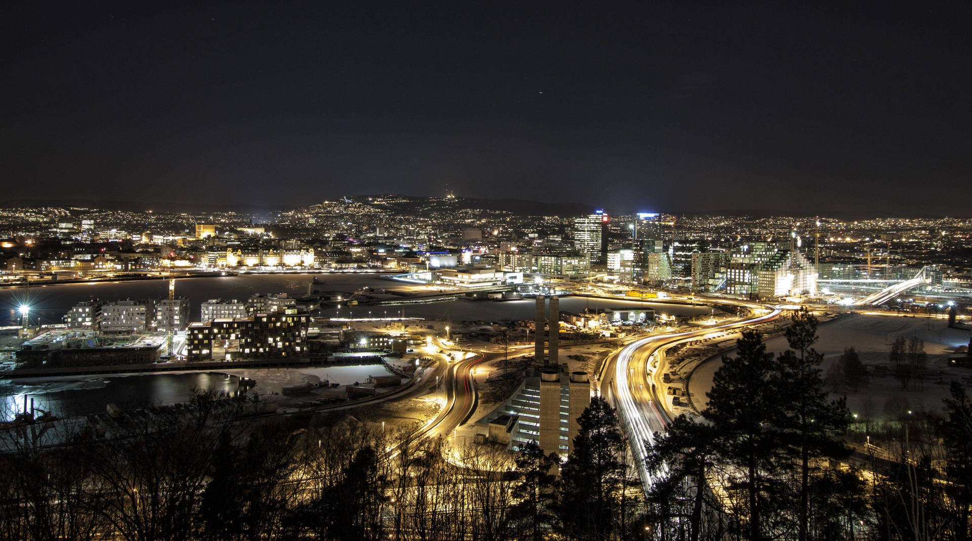 夜間城市景觀，有燈光璀璨的建築物和積雪道路，以樹木為框。