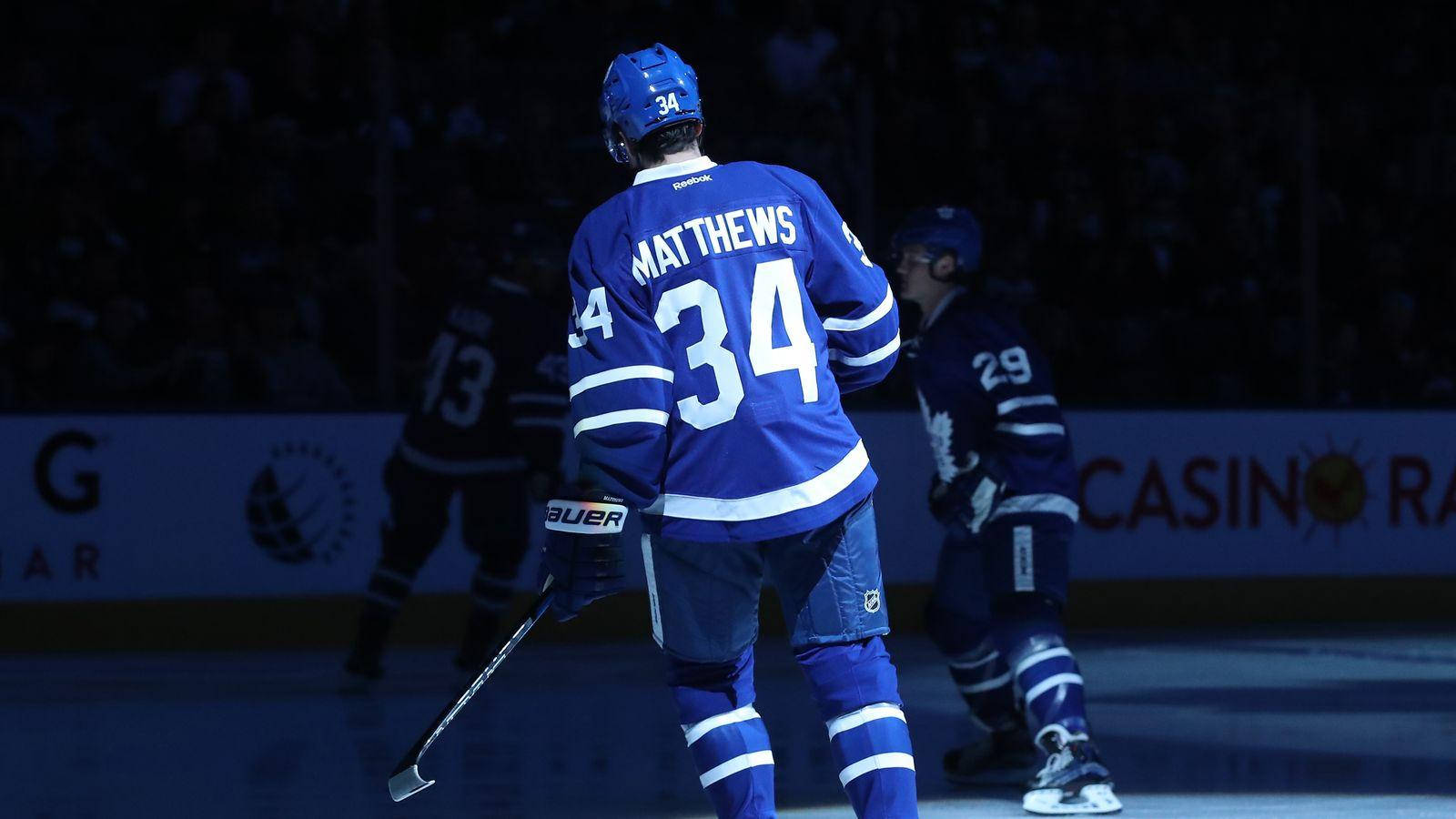 奧斯頓·馬修斯背影 壁紙 — 身穿藍色球衣的冰球運動員 Matthews 34號在比賽期間站在冰場上，場館燈光照射下。