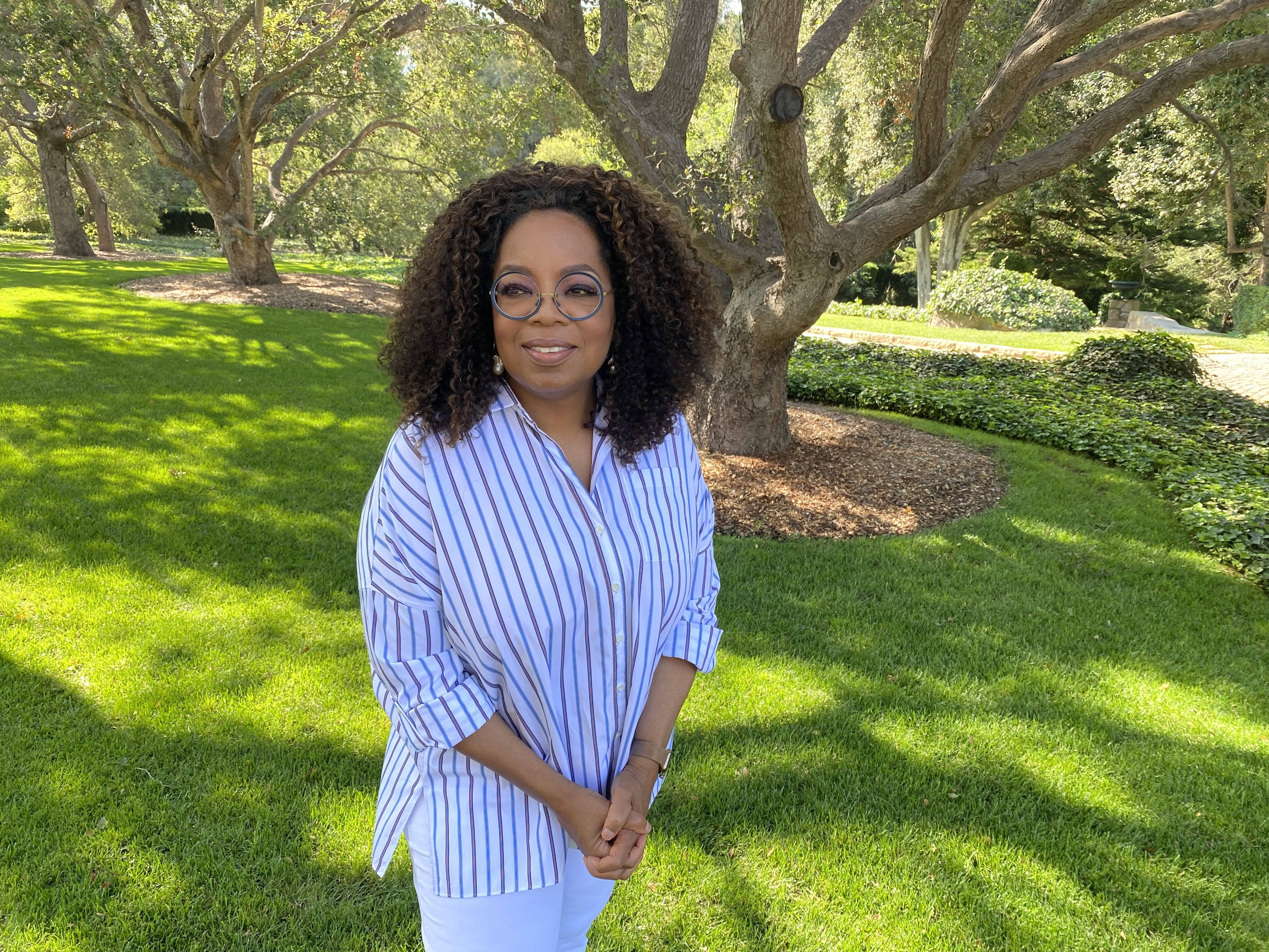 奧普拉·溫弗瑞休閒造型 壁紙 — 奧花·雲費（Oprah Winfrey）身穿條紋襯衫，站在枝葉茂密的樹下，在陽光明媚的綠色草坪上溫暖地微笑。