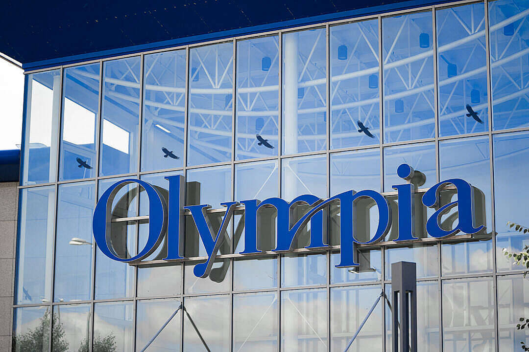現代玻璃建築物，帶有藍色Olympia標誌同反射中飛緊嘅雀仔。