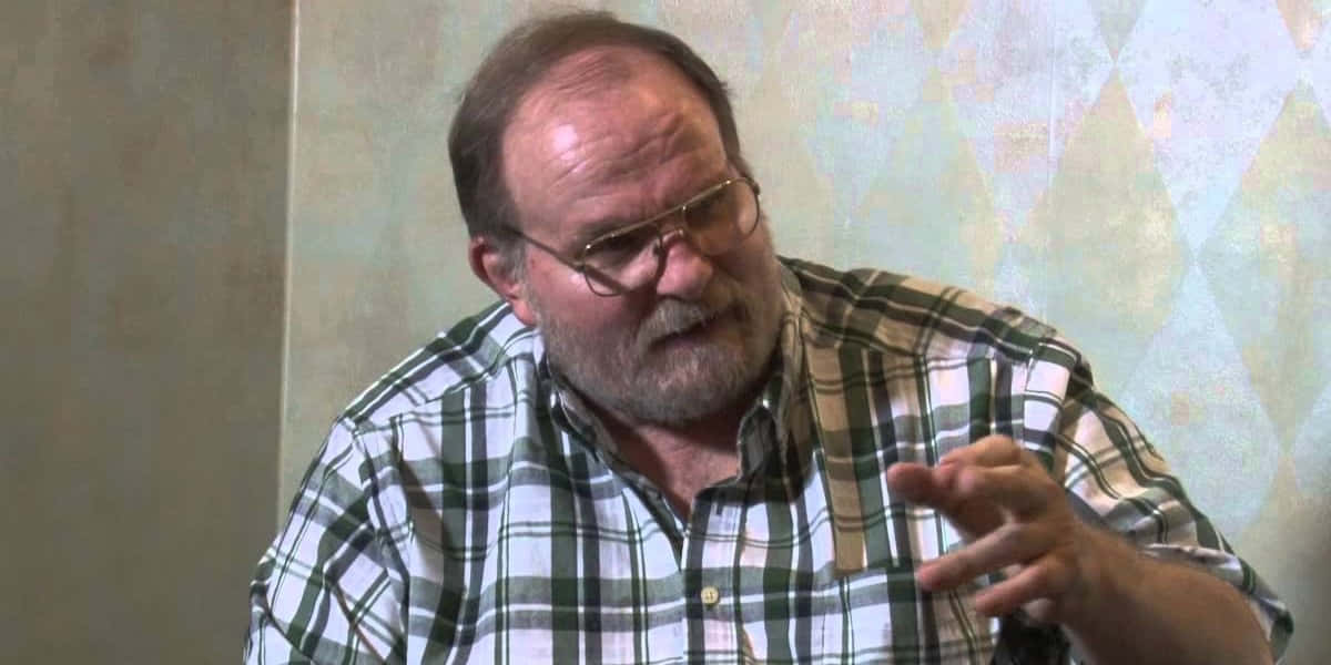 戴眼鏡留鬍鬚的中年男性在室內說話時做手勢。