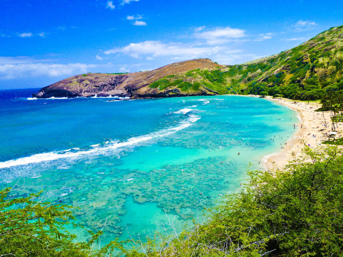 奧胡島清澈海水海灘 壁紙 — 一個充滿活力的藍綠色海灣蜿蜒環繞著沙灘，沙灘上有游泳者和日光浴者，天空湛藍，白雲點點。