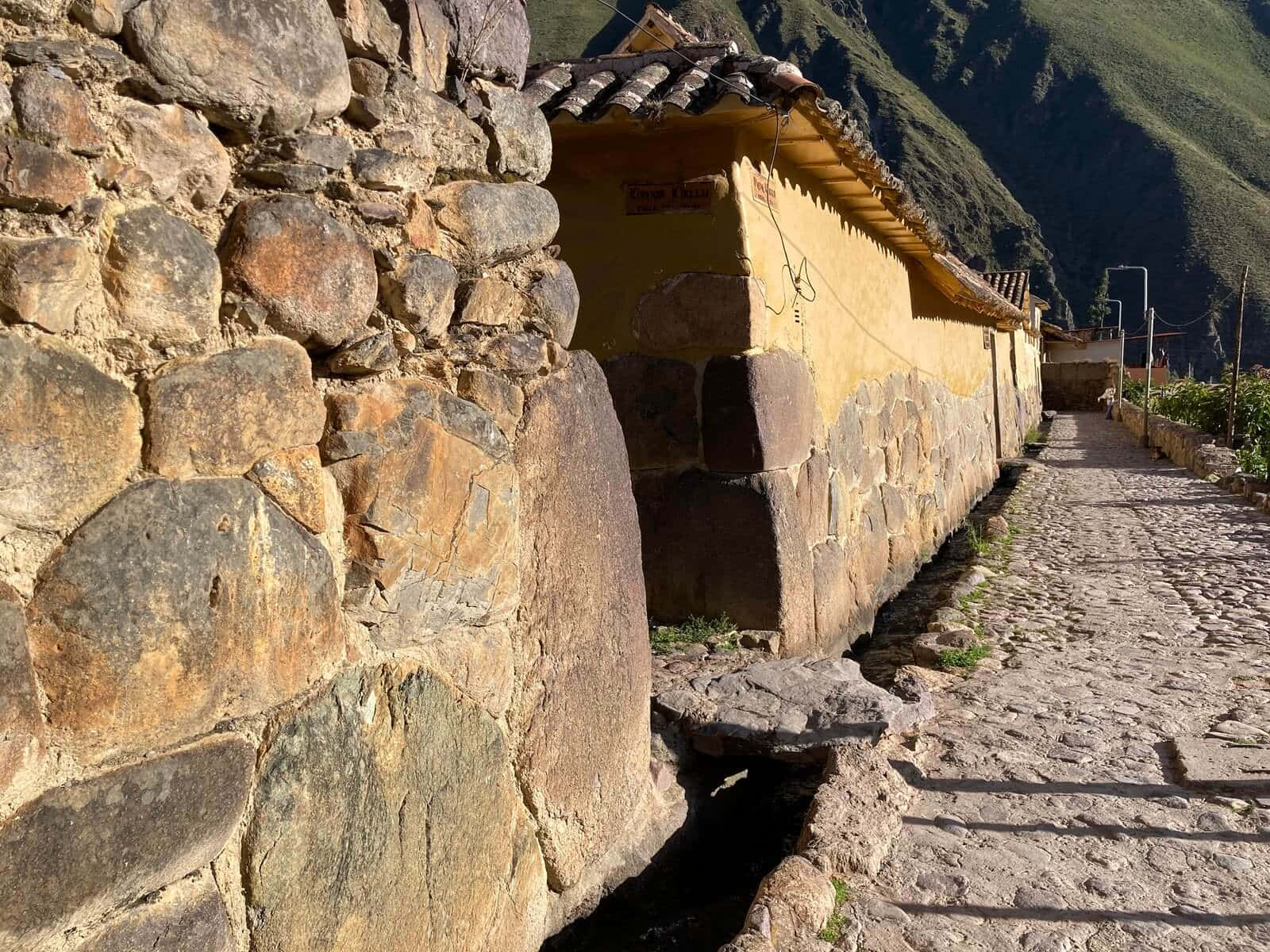 奧蘭塔坦博石階梯 壁紙 — 一條陽光普照的鵝卵石小徑蜿蜒穿過古老石牆和一座坐落在山谷中的黃色灰泥建築。