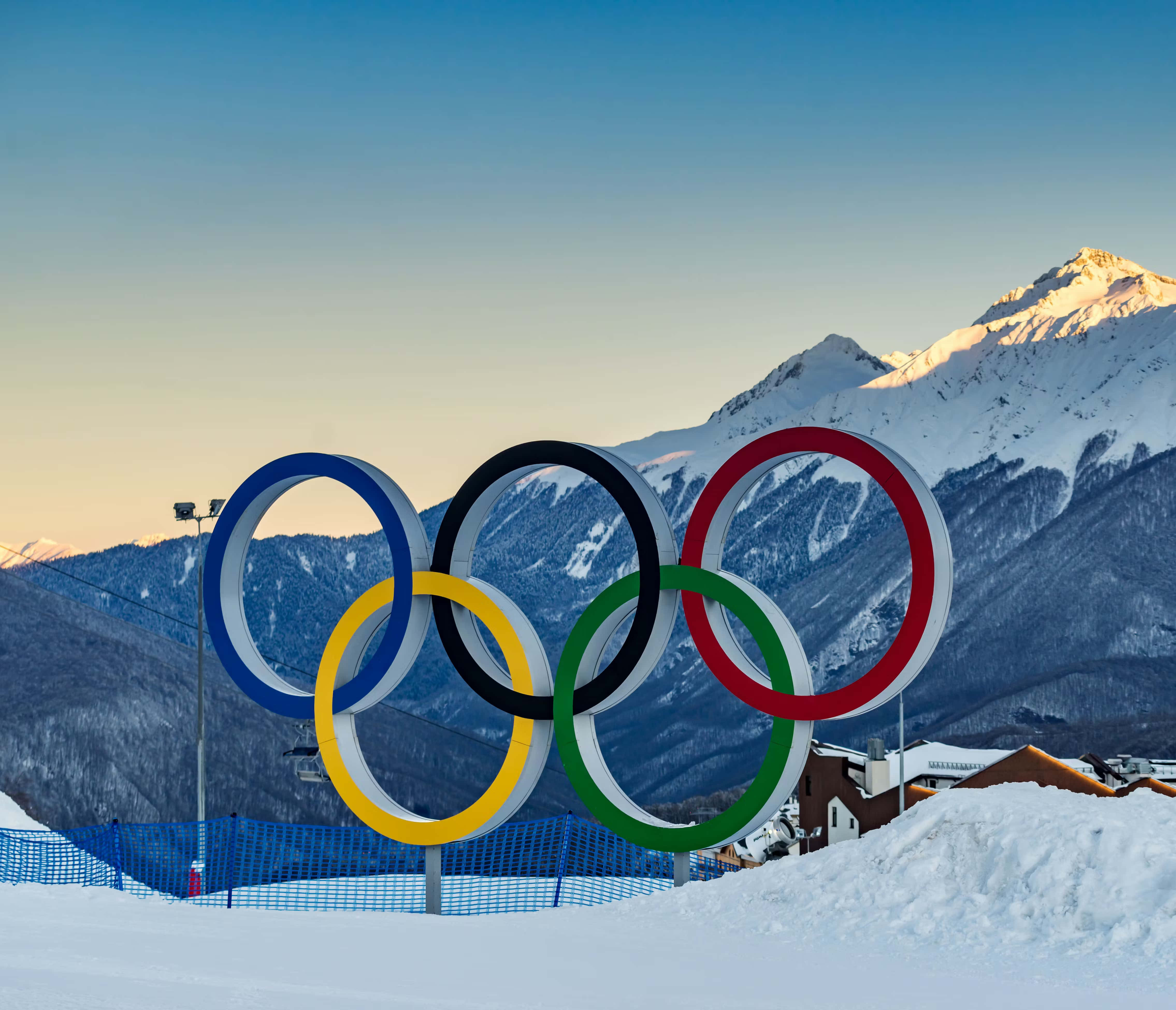 Olympics 桌布 壁紙 — 奧運五環在日出時分，在晴朗藍天下，聳立在積雪覆蓋的山脈前。
