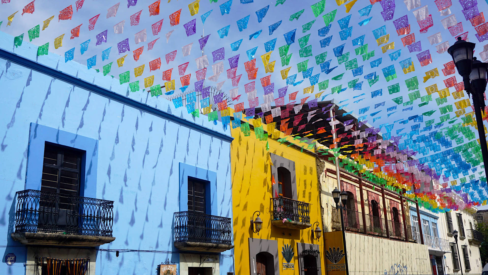 色彩繽紛的 papel picado 橫幅掛在節日街道上，街道上有藍色和黃色的建築。