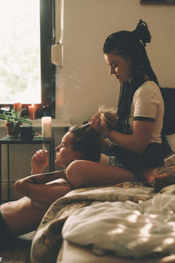 兩名女子在臥室中，一人為另一人編辮子，同時手持香煙。