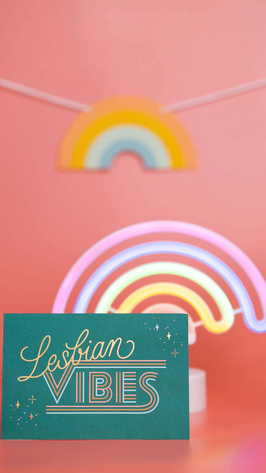 一張青綠色卡片，帶有「Lesbian Vibes」文字，放在發光的霓虹彩虹前。