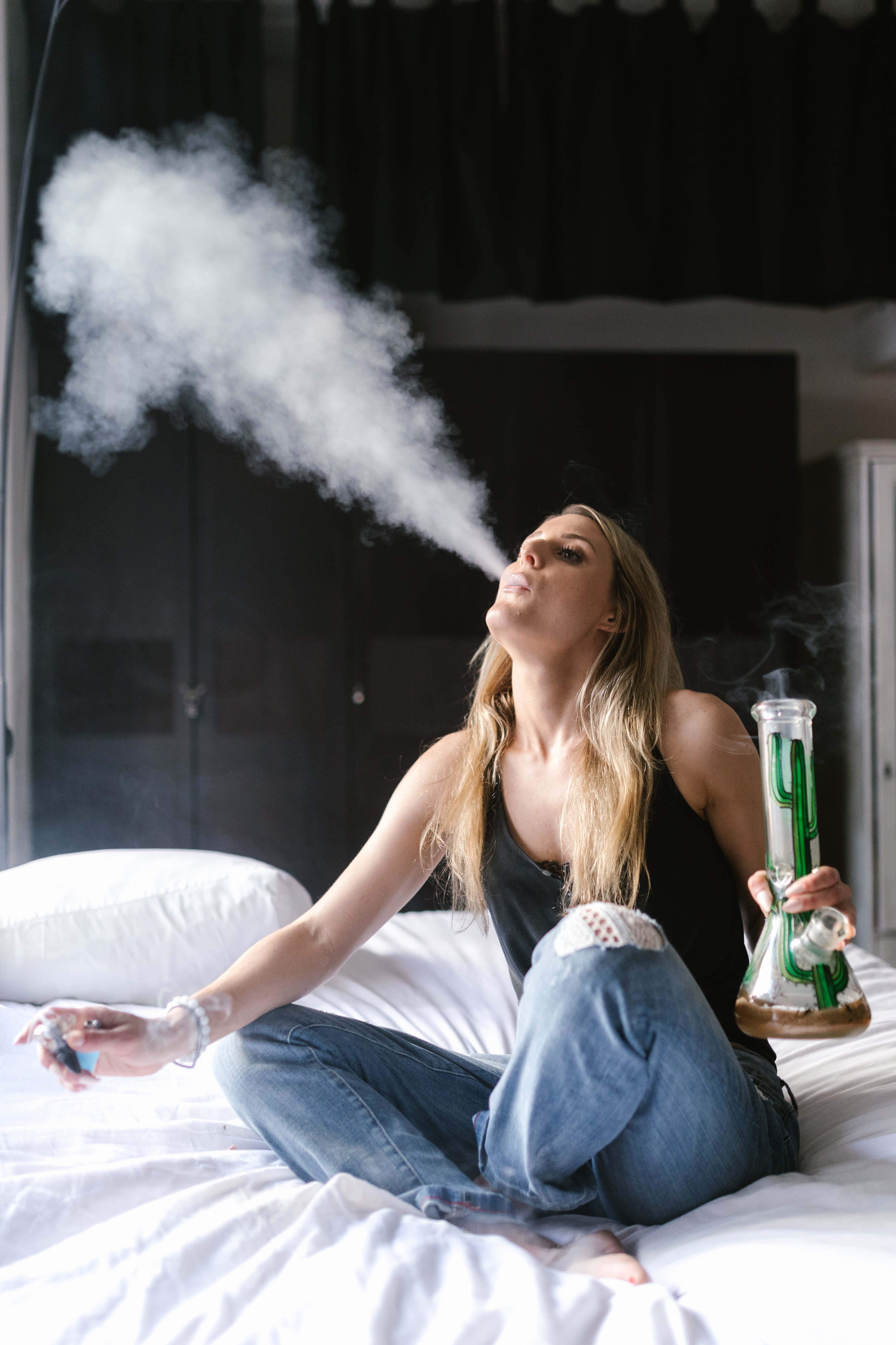 女子在床上使用水煙壺搭配煙草 壁紙 — 一名女性盤腿坐在床上，呼出一團濃煙，同時手持玻璃水煙槍。