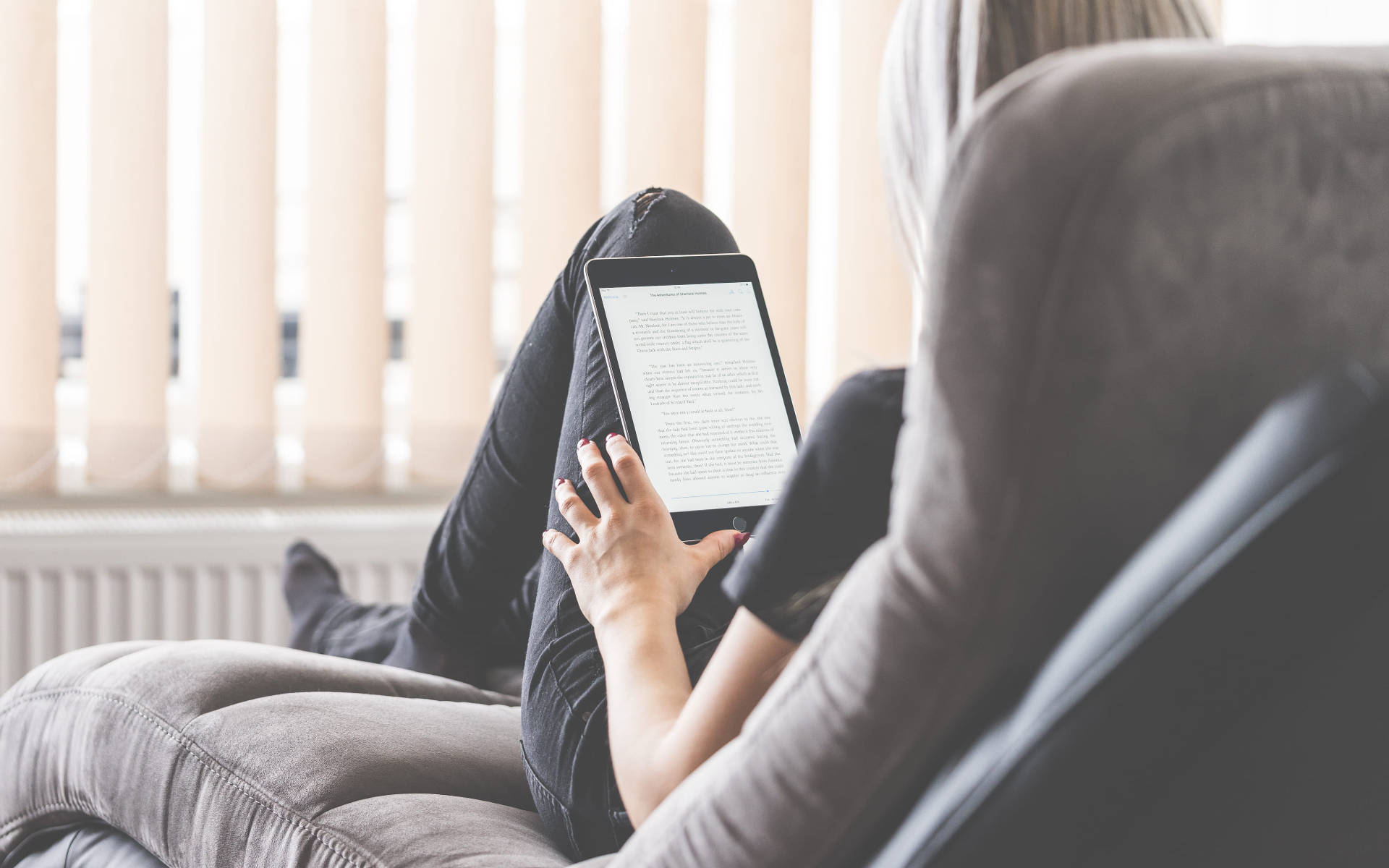 女子在沙發上閱讀電子書 壁紙 — 一個人舒適地躺在灰色扶手椅上，用平板電腦看書，柔和自然光透過垂直百葉簾照射進來。