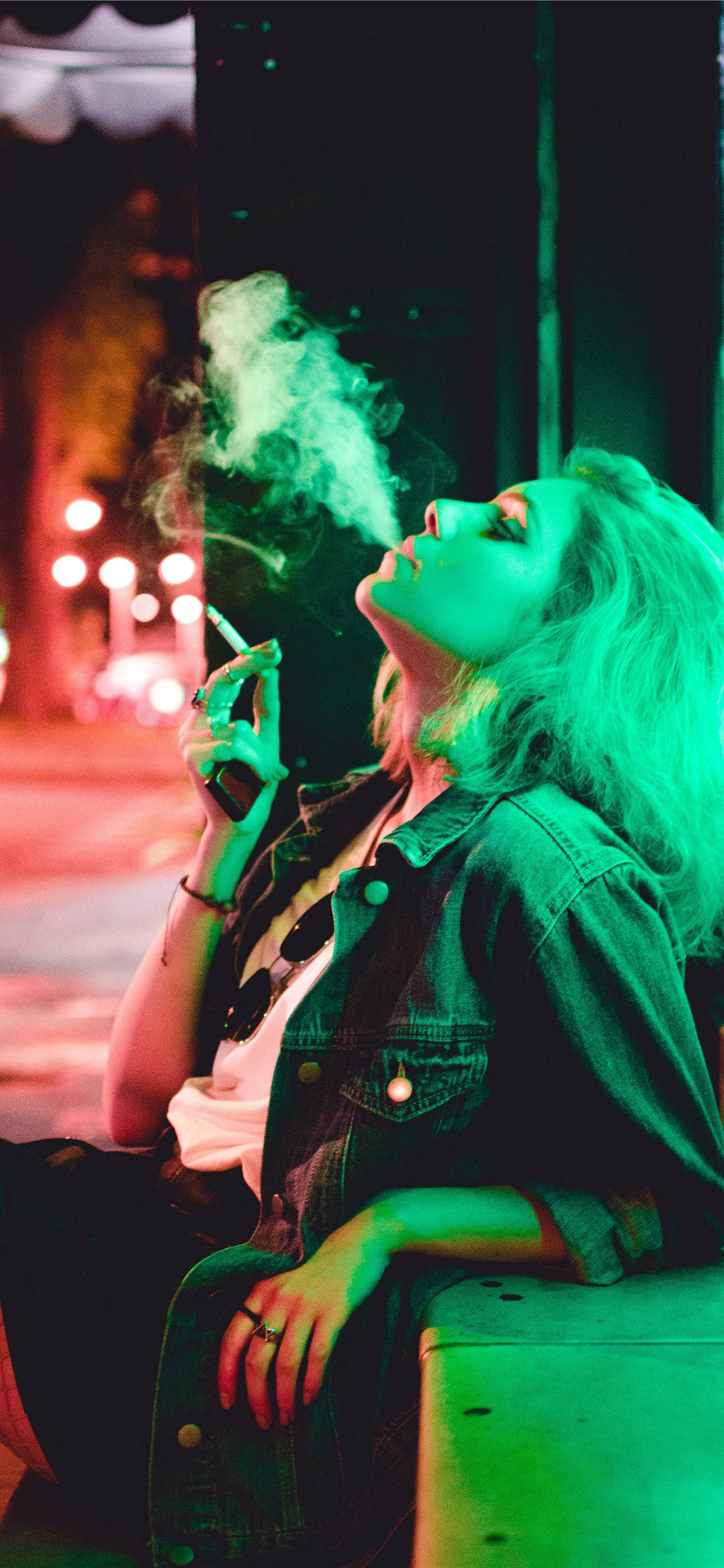 女子抽菸暗色霓虹iPhone壁紙 壁紙 — 一位金髮女子在夜晚坐在窗台上，在霓虹綠光和紅光下呼出煙霧。