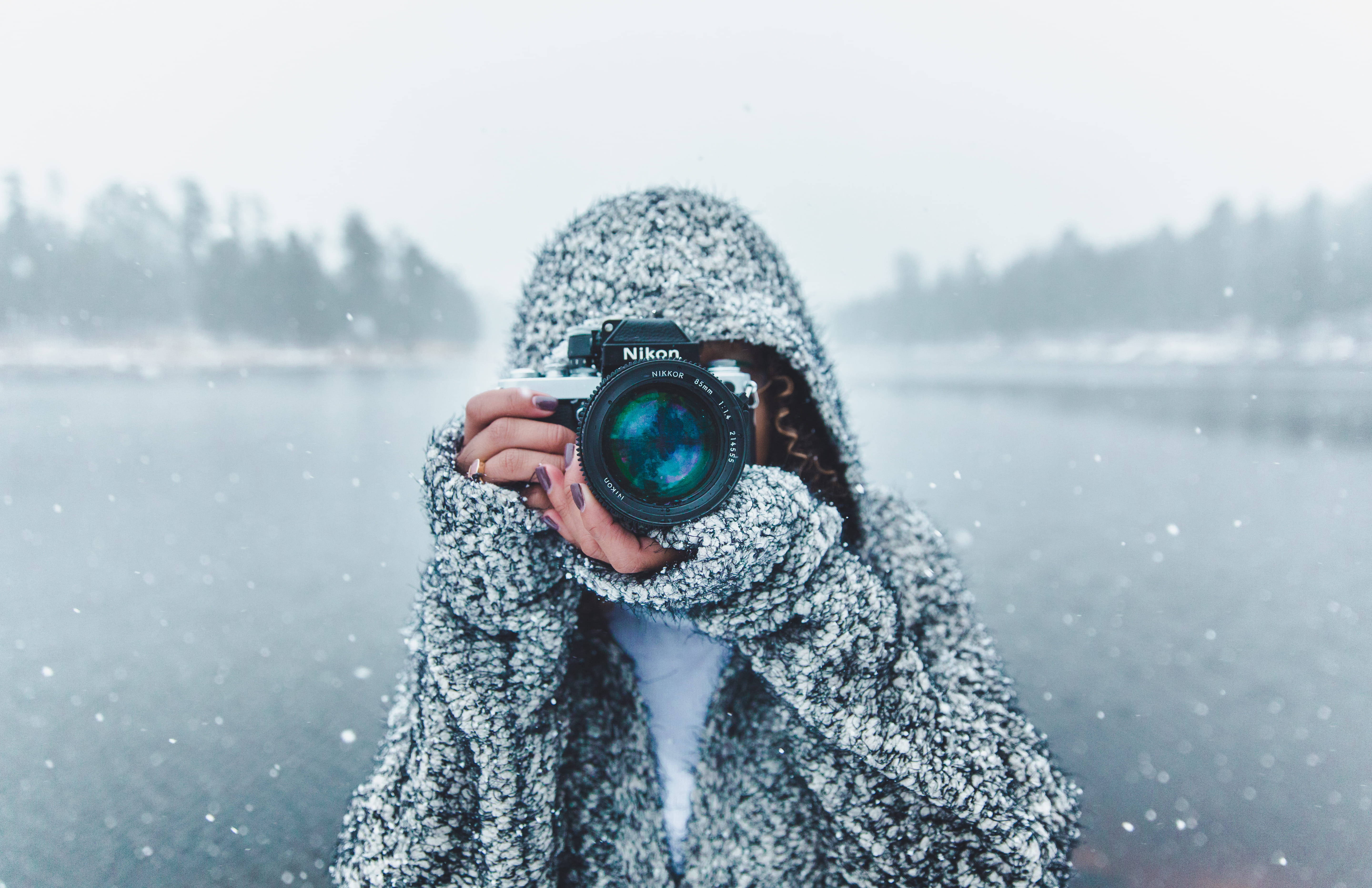 女子於極寒天氣中 壁紙 — 一個穿著質感連帽外套的人，在雪白模糊的冬季風景中將相機舉到臉前。