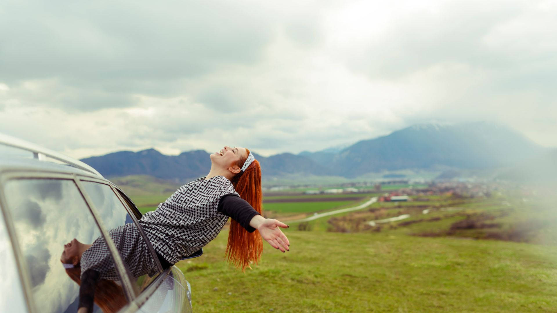 女子旅程 壁紙 — 一位紅髮的快樂女士從車窗探出身子，張開雙臂，在陰天背景下，對著起伏的綠色山丘和遠山大笑。