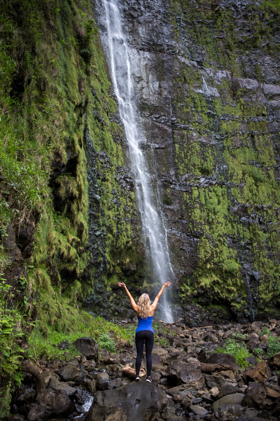 女子森林中享受自由 壁紙 — 一位高舉雙臂的女性敬畏地站在一道從長滿苔蘚的懸崖上傾瀉而下的高瀑布前。