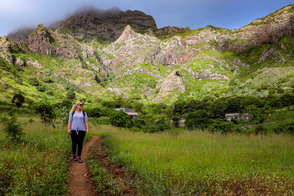 一名女子在泥路上徒步穿過綠色田野，背景有山脈。