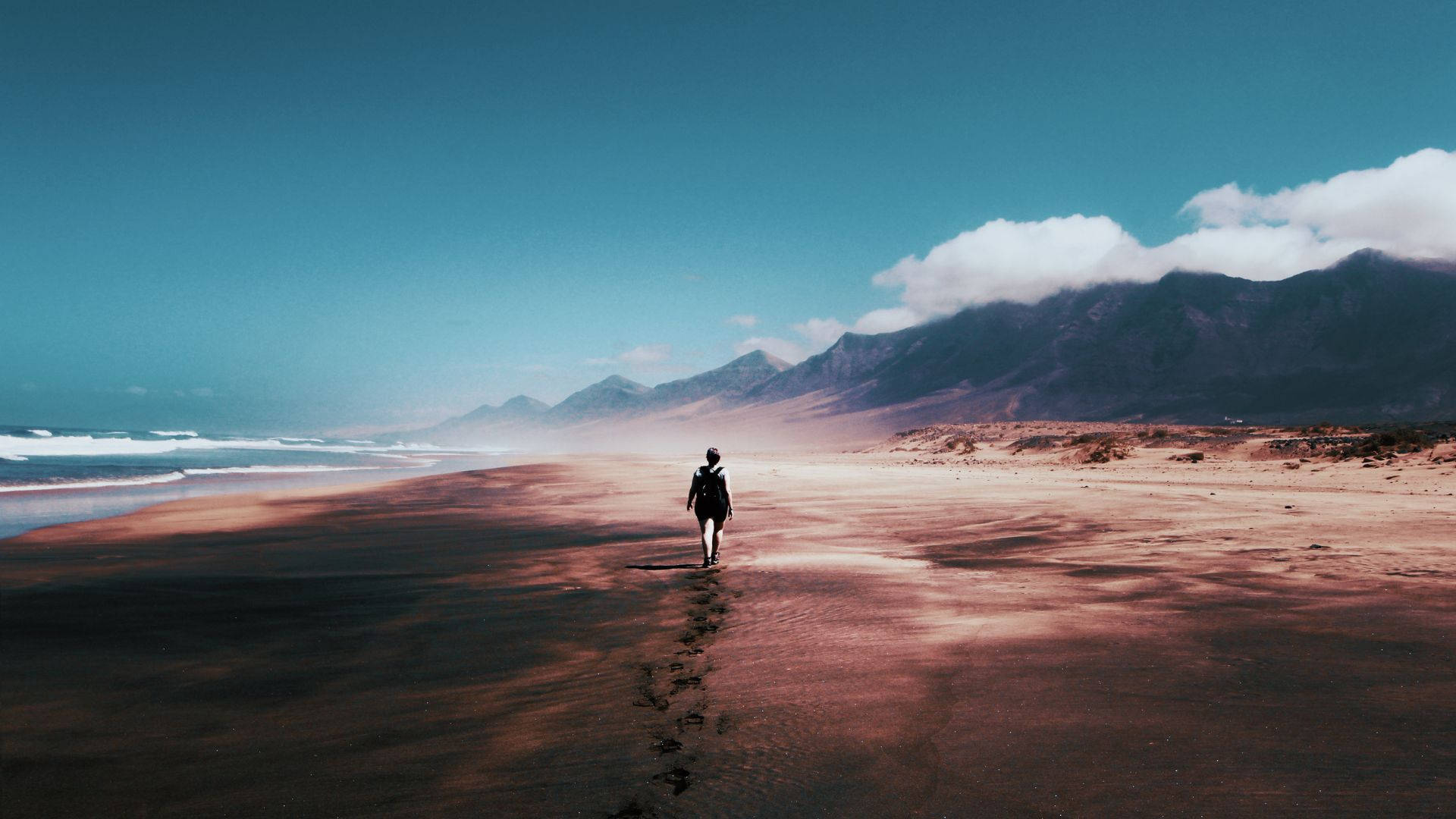 女子獨自行走海灘的旅程 壁紙 — 一個孤獨的身影沿著廣闊的粉紅色沙灘走向遠山，天空湛藍清澈，白雲點綴其間。