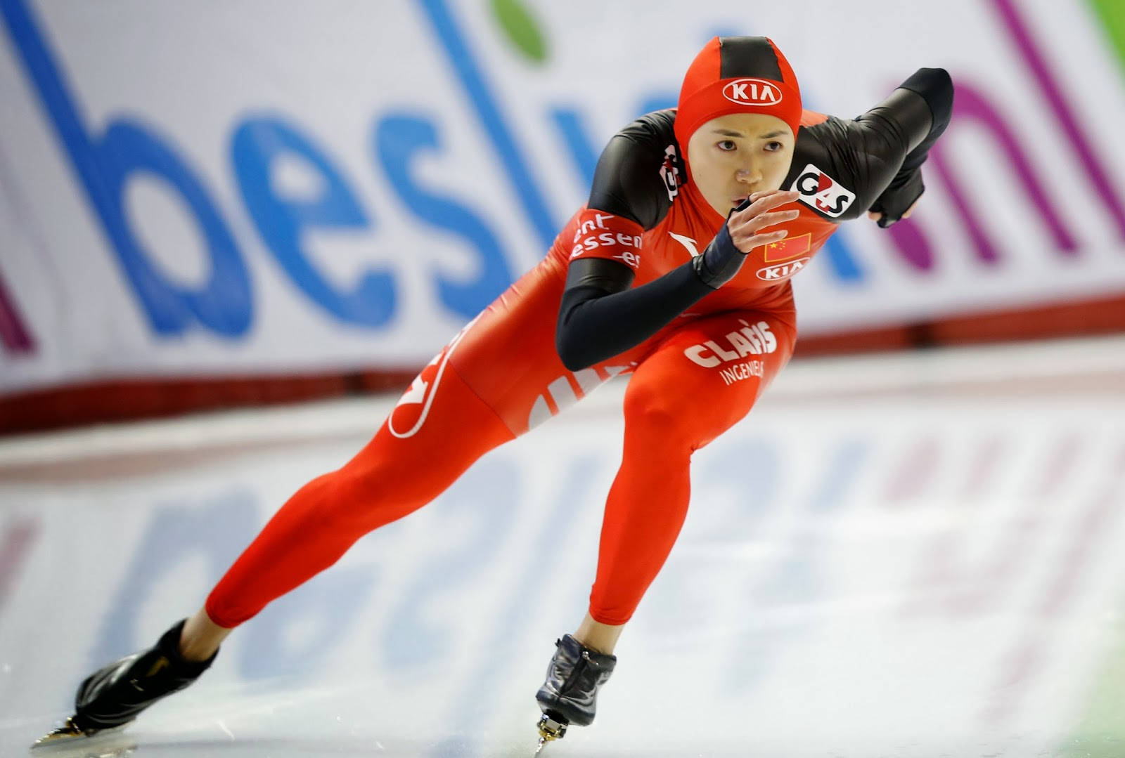 女子速滑 壁紙 — 一名身穿紅黑套裝的女子速度滑冰選手在室內溜冰場上傾身過彎，身後有模糊的贊助商橫幅。