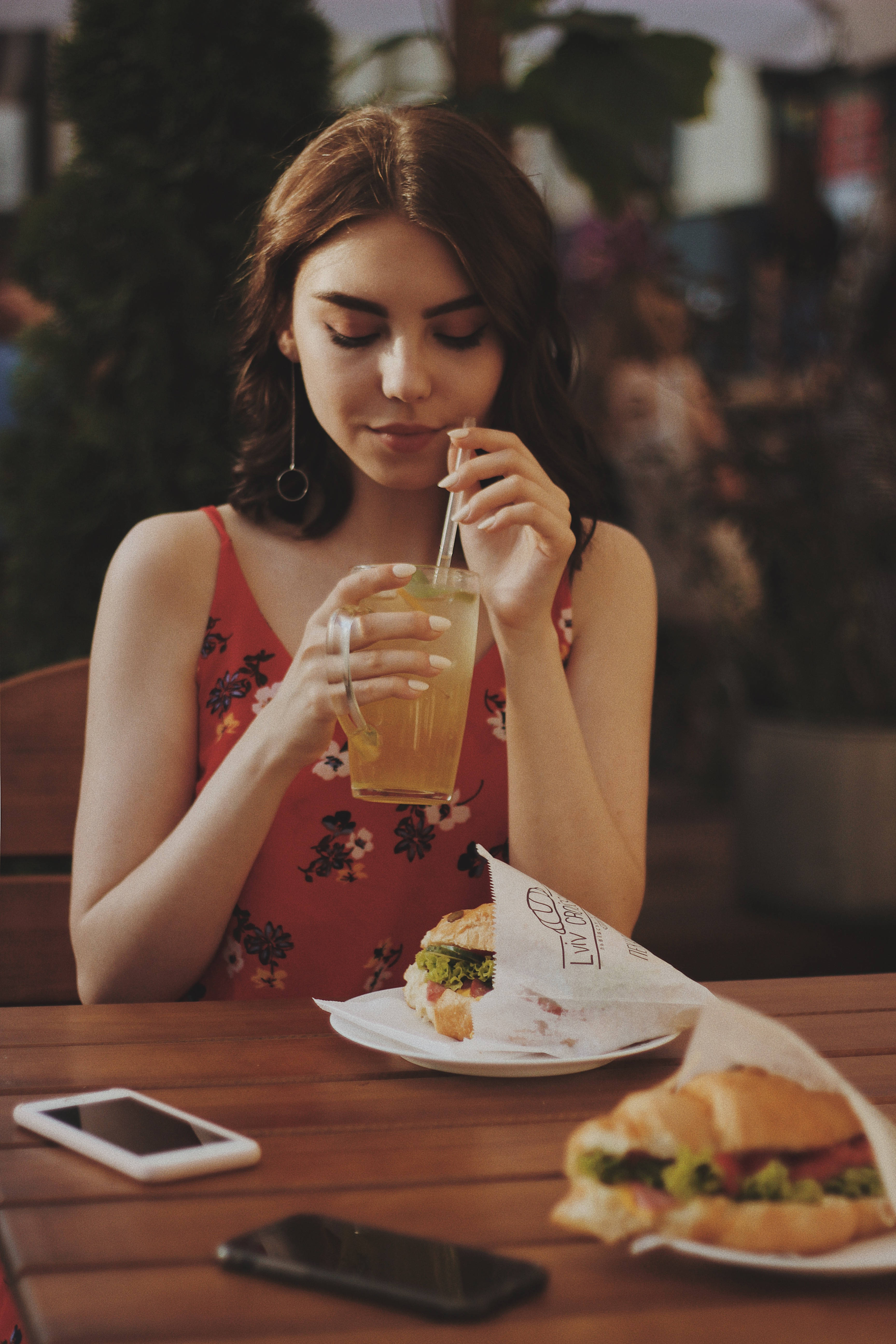 女子飲冰茶美食 iPhone 壁紙 — 一位年輕女性在戶外咖啡桌旁啜飲清涼飲品，旁邊放著三文治和電話。