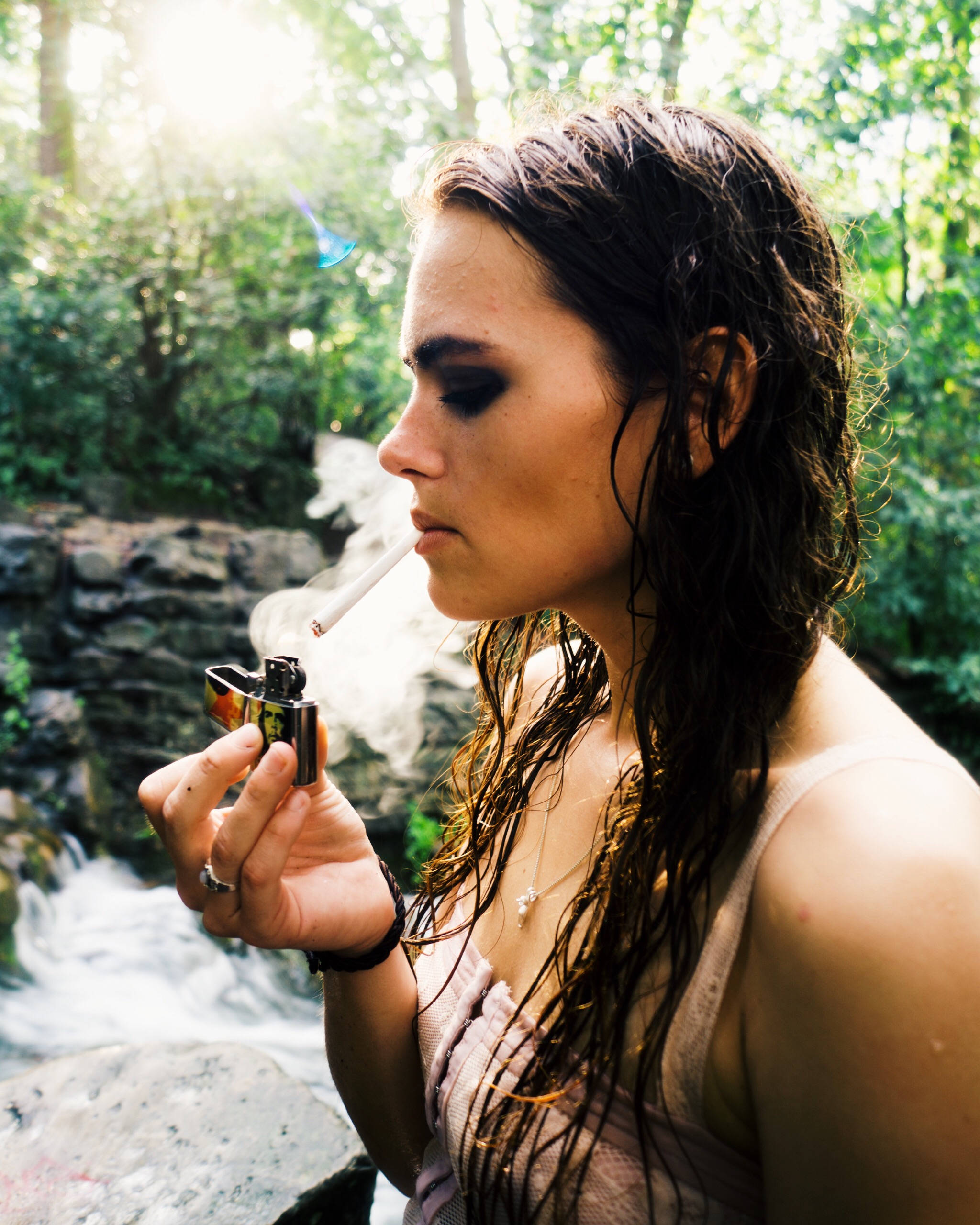 女子點燃大麻煙卷 壁紙 — 一位濕髮女性在陽光明媚的森林溪流旁抽煙，手持一個色彩繽紛的打火機。