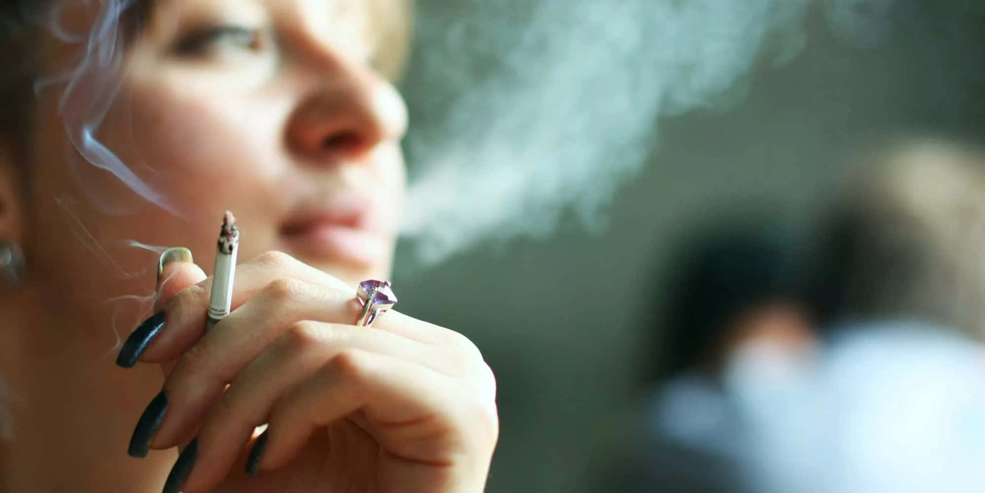 女孩吸菸的照片 壁紙 — 一名女性呼出煙霧，手中拿著一支香煙，手指塗有深色指甲油並戴著紫色戒指。