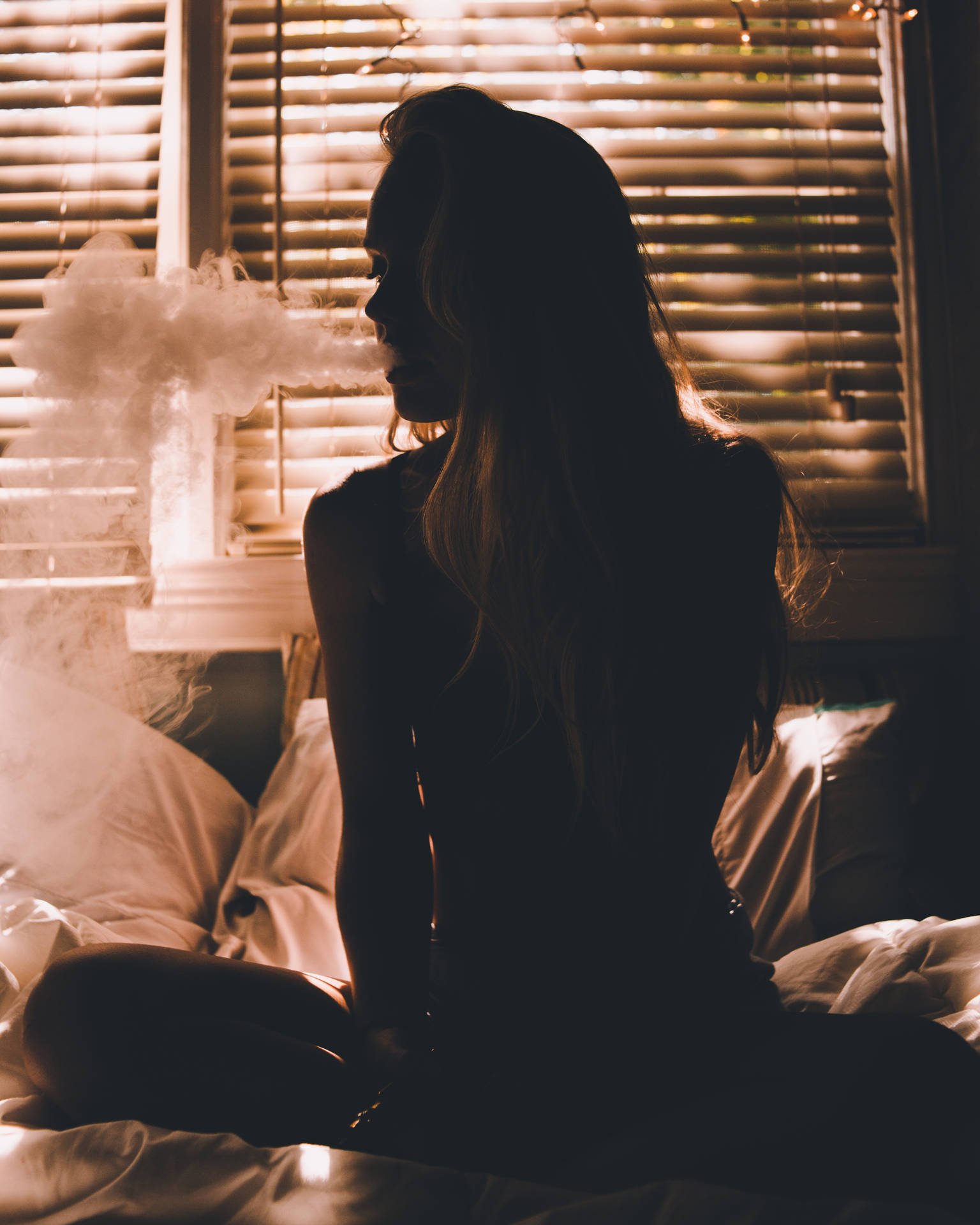 女人坐在床上，在昏暗的房間裡透過百葉窗吐煙