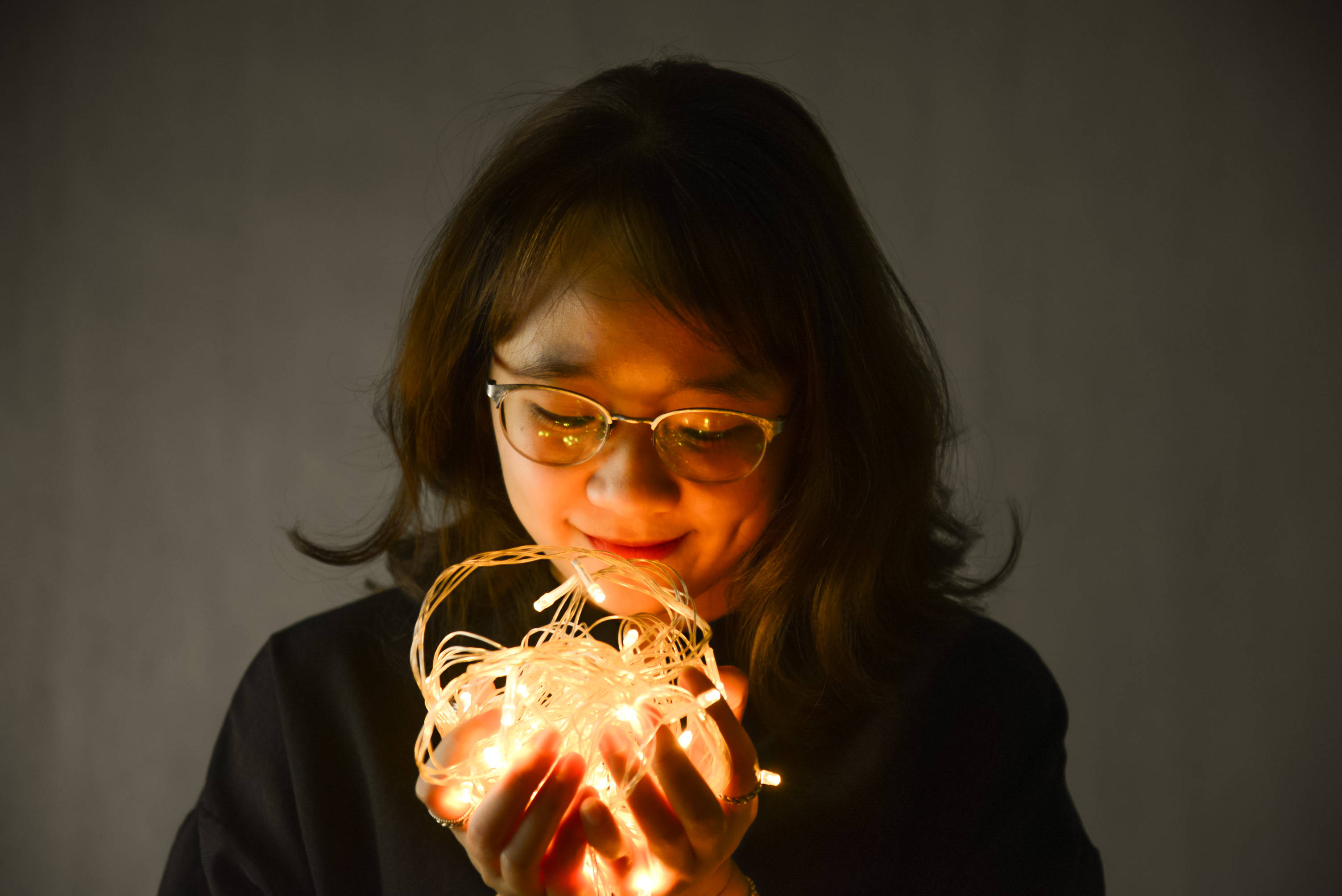 女孩手持精美聖誕燈飾 壁紙 — 一位戴眼鏡的年輕女子輕輕懷抱著發光的仙女燈，她的臉龐在柔和的灰色背景下被輕輕照亮。