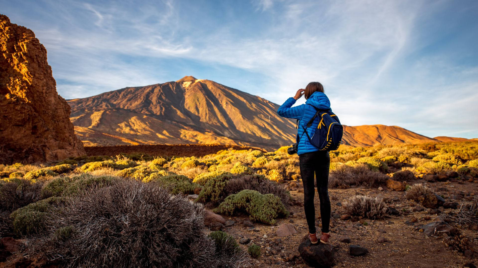 女孩攀登泰德火山之旅 壁紙 — 一位身穿藍色外套的遠足者站在岩石小徑上，凝視著在明亮藍天和縷縷白雲下的雄偉山脈。