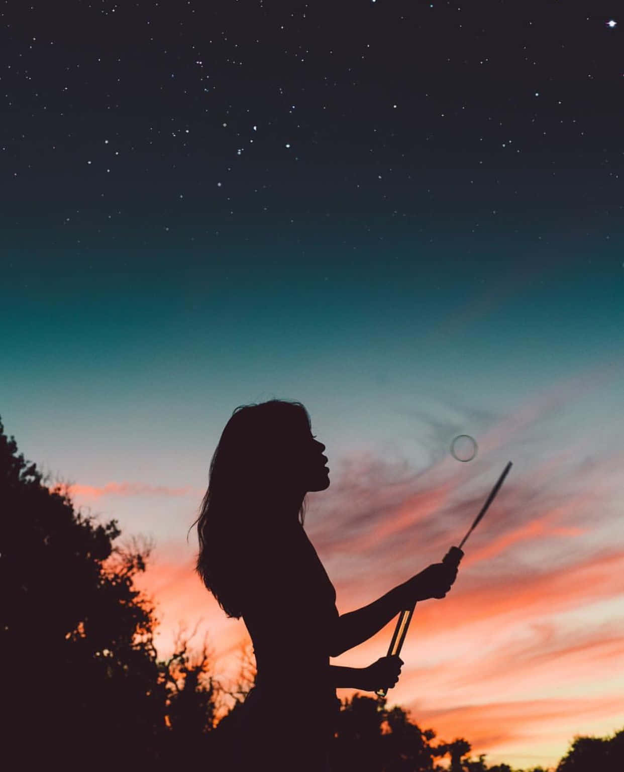 女孩立於夜色陰影中 壁紙 — 一個女人的剪影，在黃昏的天空下吹著泡泡，天空上有橙色、粉紅色和青綠色的星星條紋。