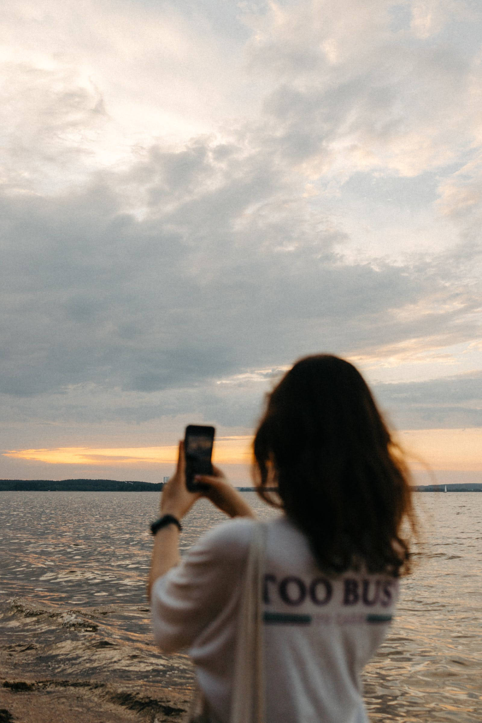 女孩自然 iPhone 背景 壁紙 — 一名深色頭髮的人在海濱拍攝日落，天空多雲。