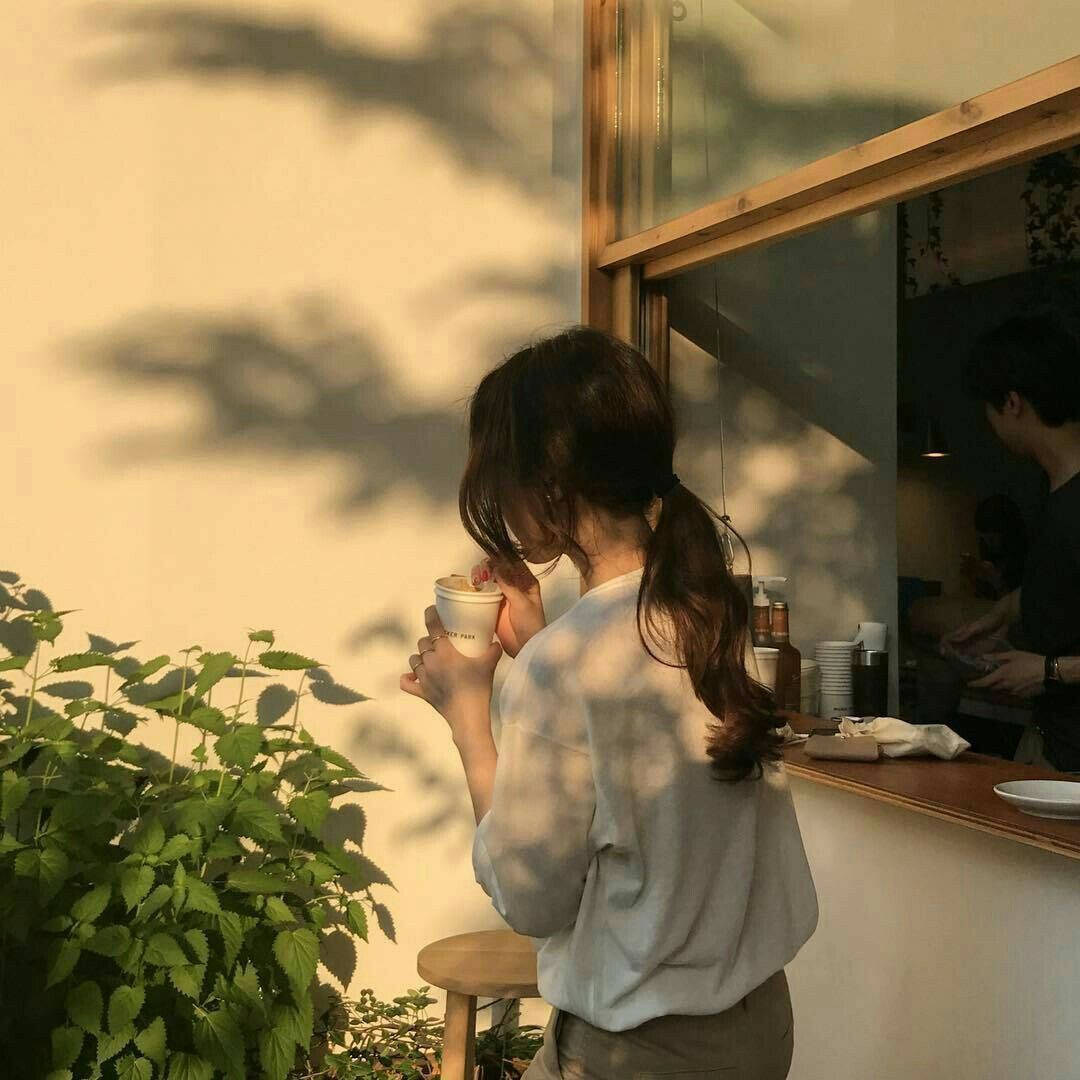 女士在陽光明媚的咖啡館喝咖啡，附近有樹影和綠色植物。