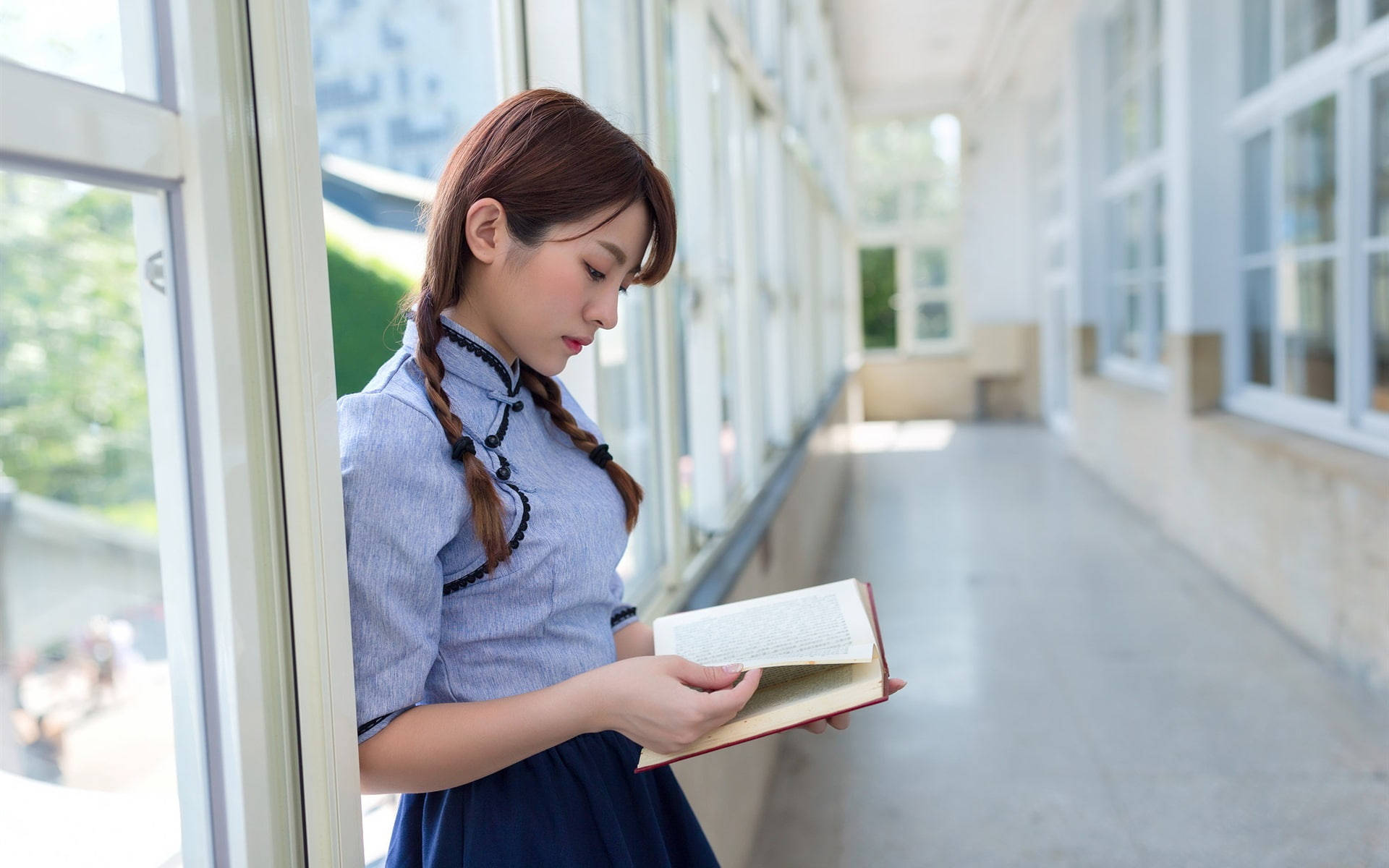 女學生走廊閱讀 壁紙 — 一位身穿藍色校服的年輕女子在明亮的學校走廊裡靠著窗戶看書。