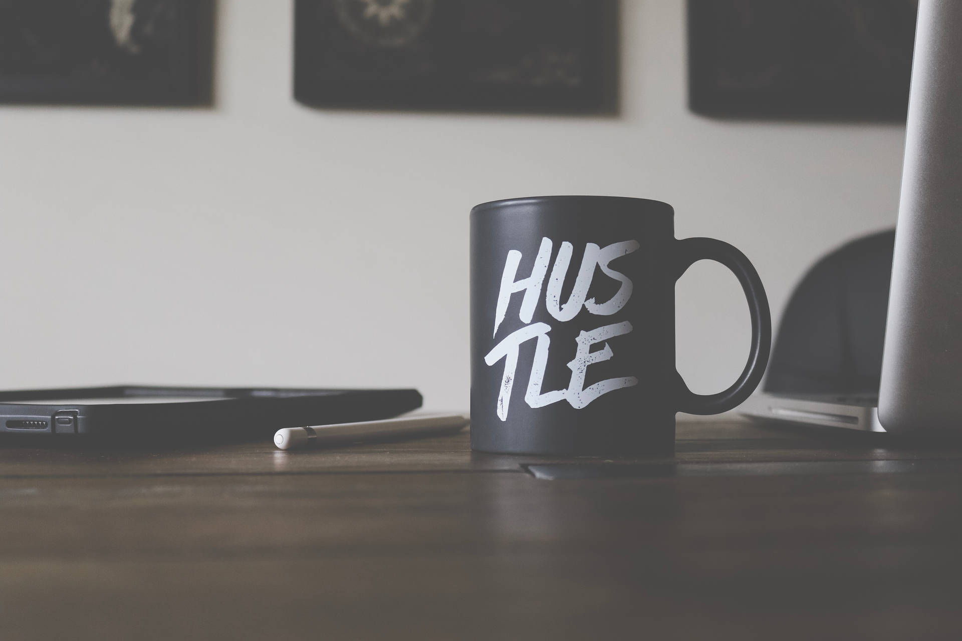 黑色馬克杯帶白色 hustle 文字，木桌上有手提電腦和平板電腦。