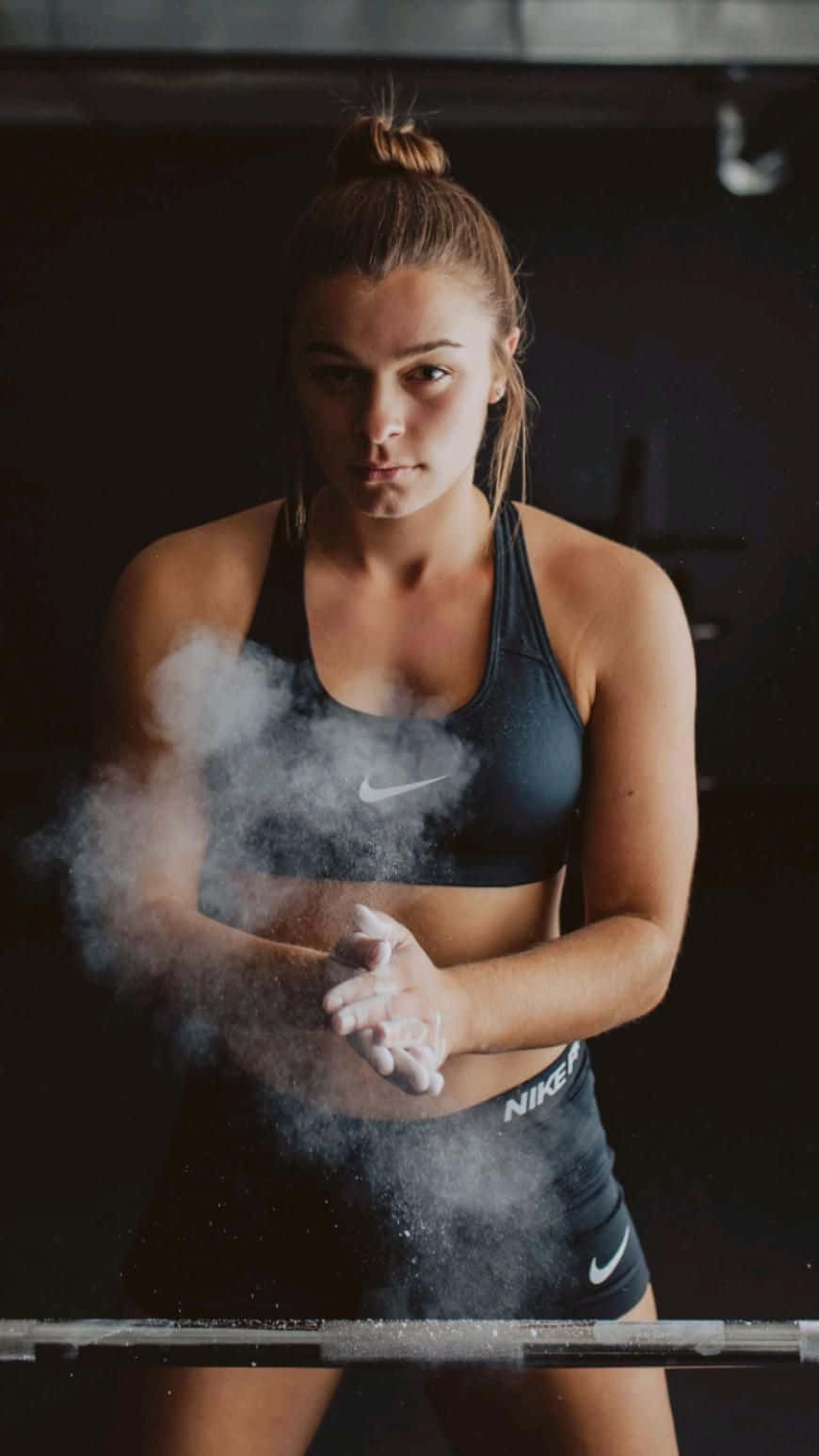 女性在槓鈴上塗粉 壁紙 — 一位專注的女運動員穿著深色Nike裝備，在昏暗的健身房中舉起槓鈴前，在手上塗抹粉筆灰。