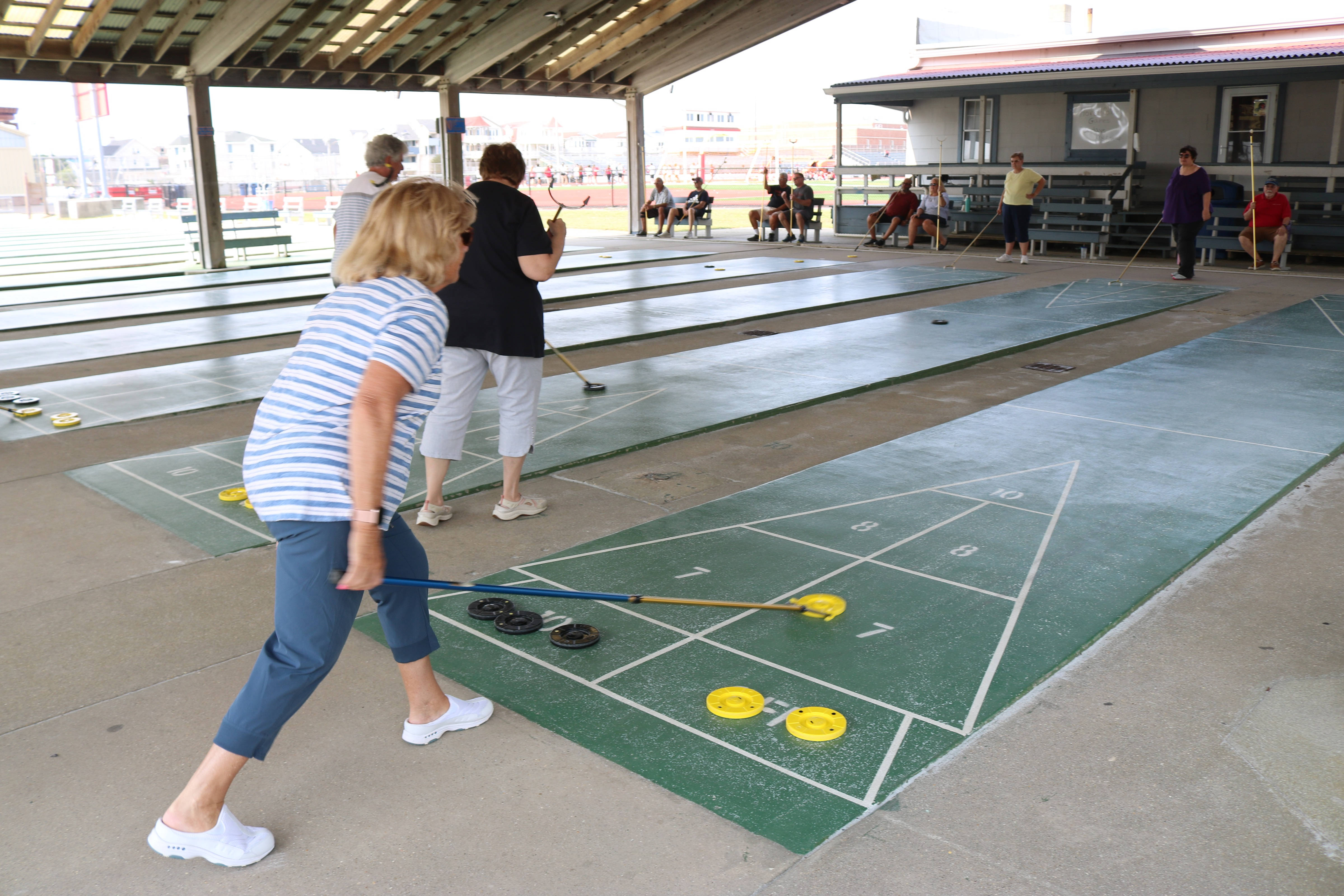 女性在球場上玩 Shuffleboard 壁紙 — 一名身穿條紋襯衫的女子在有蓋涼亭下的綠色沙狐球場上滾動黃色圓盤，觀眾在旁觀看。