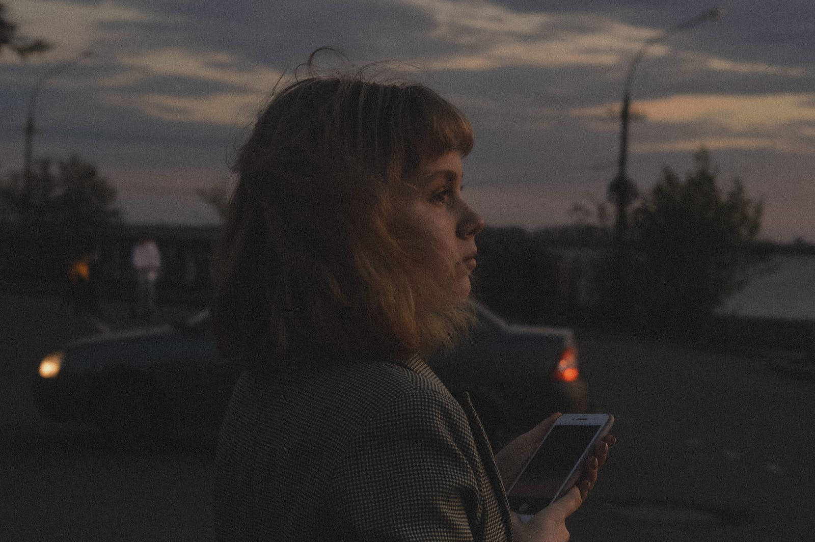 女性手持黑色iPhone 壁紙 — 一位短髮女性在黃昏時分站在寧靜的街道上，手持電話，身後有汽車駛過。