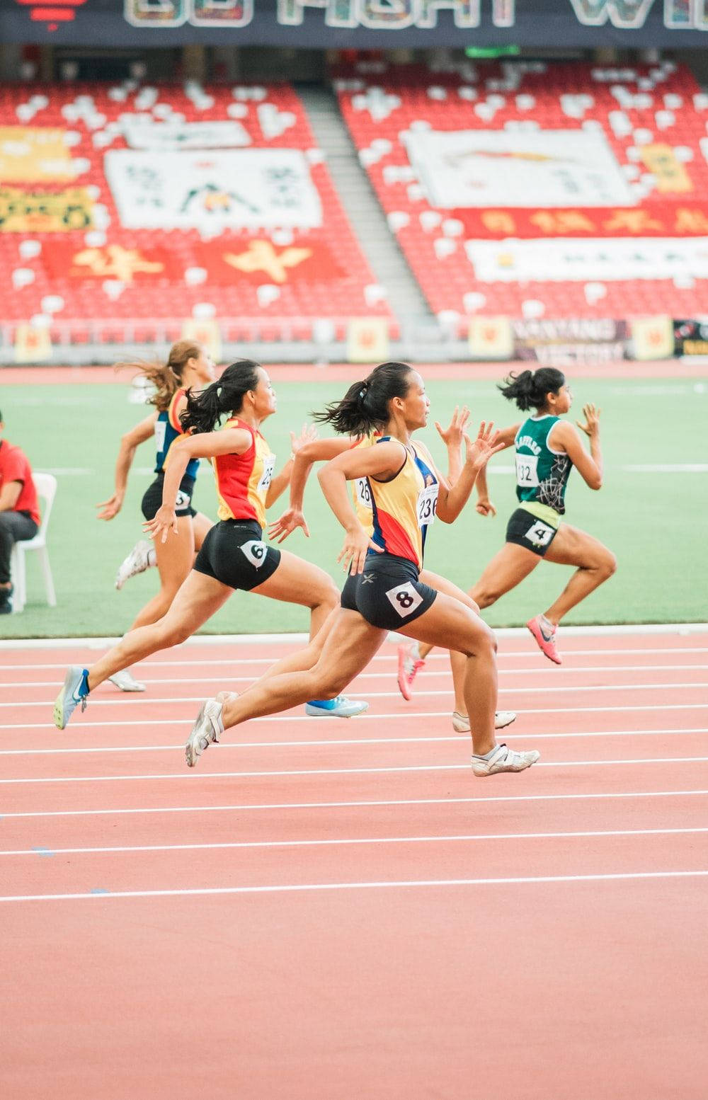 運動 桌布 壁紙 — 女性短跑選手在紅色跑道上激烈比賽，背景有模糊的動作和體育場座位。