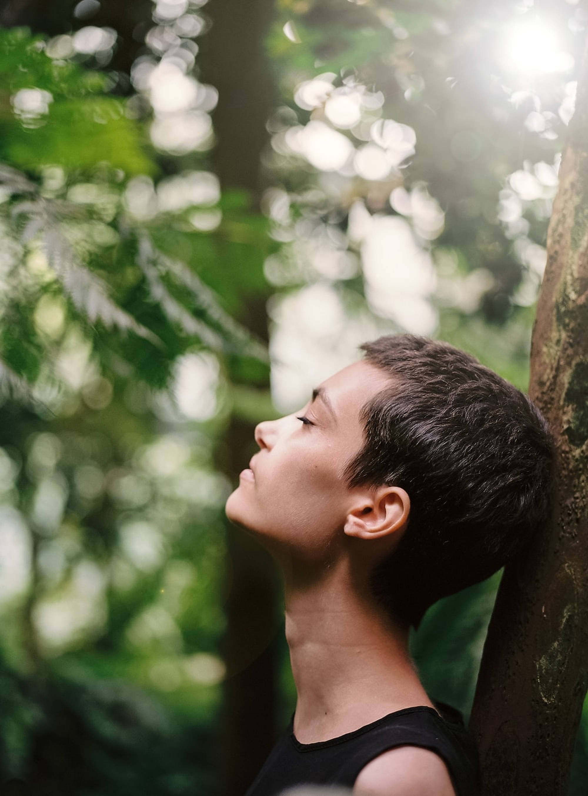 女性靜心呼吸 壁紙 — 一個短黑髮的人靠在一棵陽光明媚的森林中的樹上，閉著眼睛，享受著透過綠葉過濾的斑駁光線。