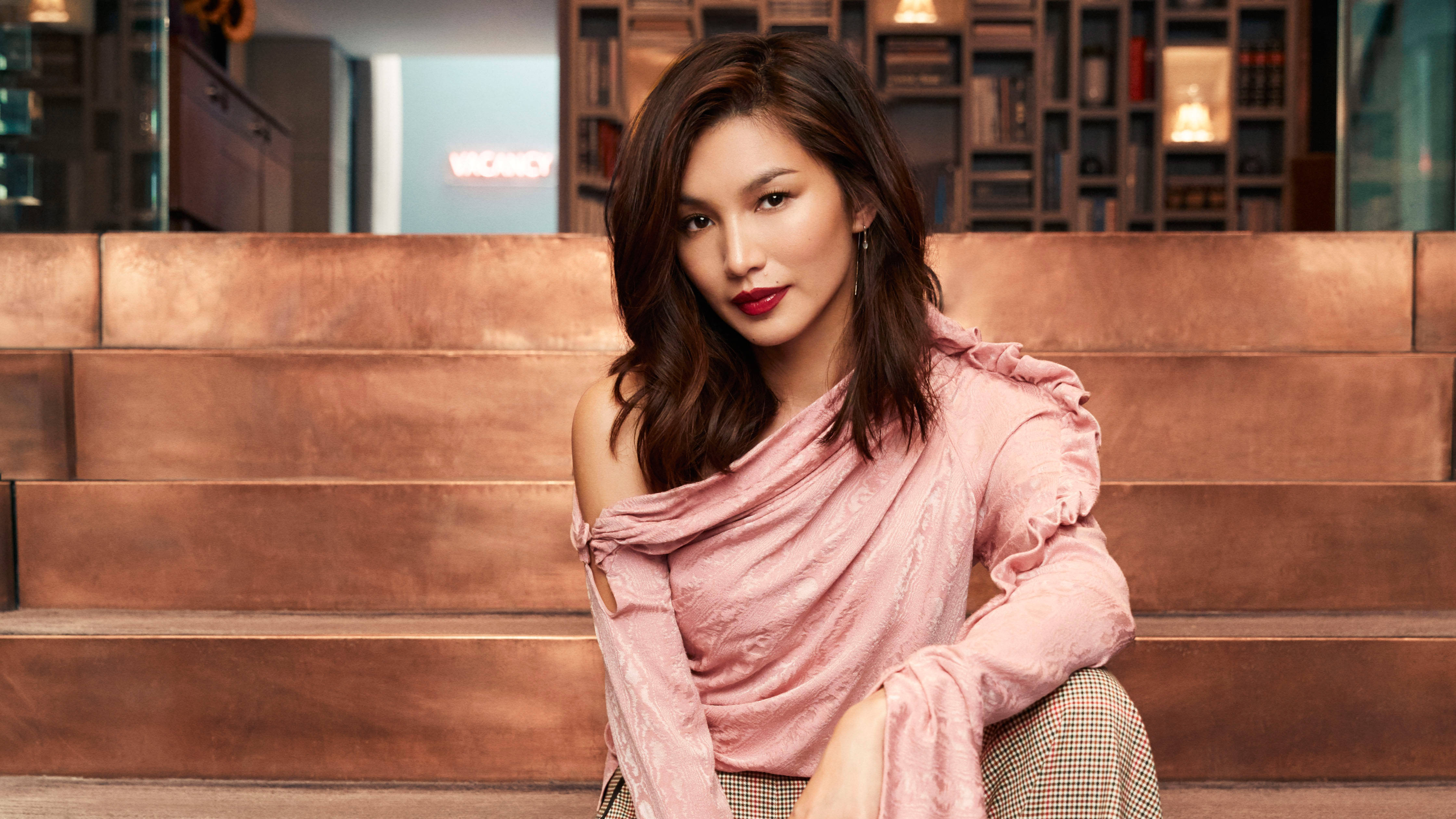 女演員嘉瑪·陳 壁紙 — 一位深色頭髮、塗著紅色唇膏的女性自信地坐在現代室內銅色調的台階上，身後有書架。
