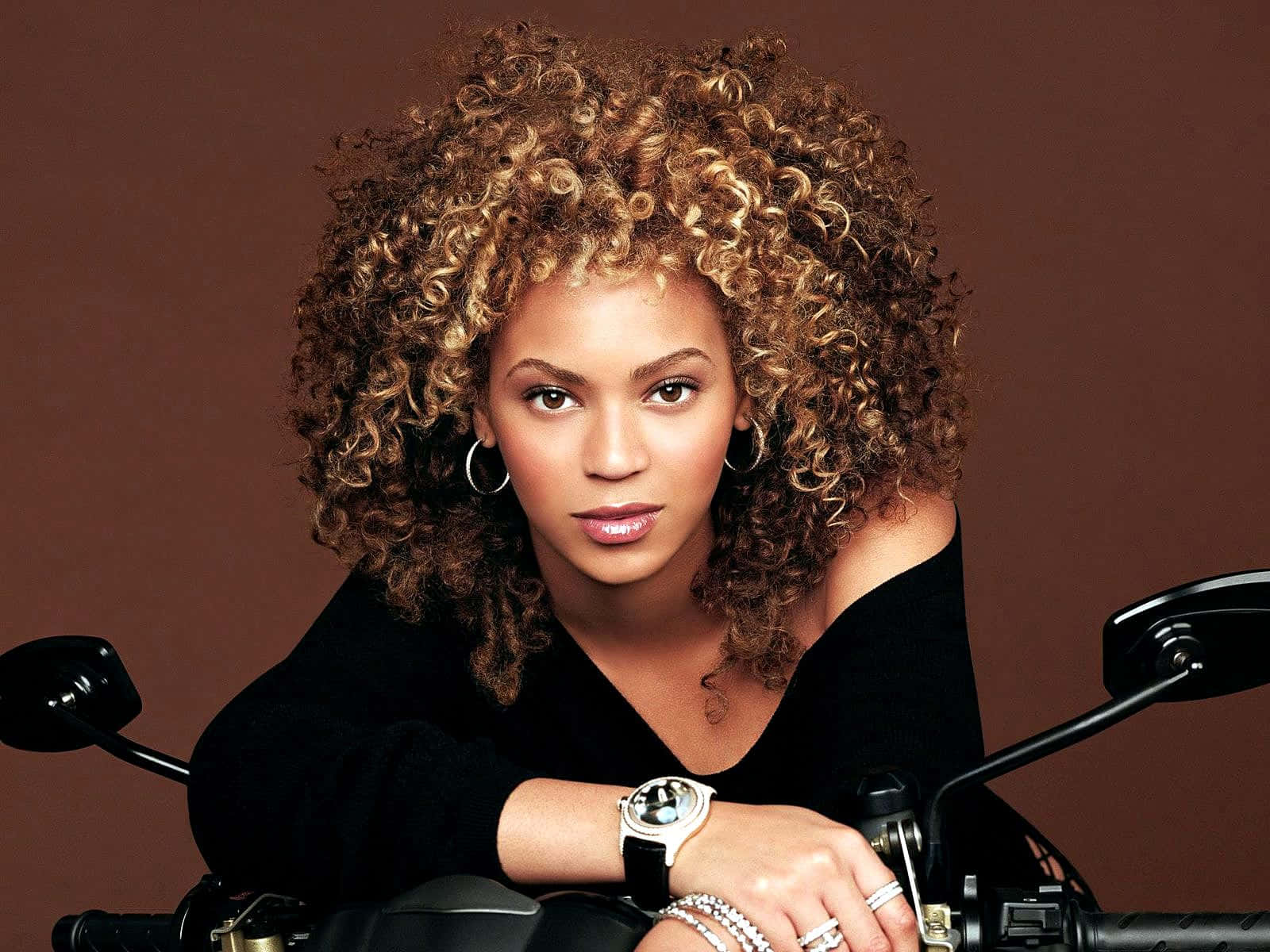 女王碧昂絲 壁紙 — Beyoncé 擁有一頭濃密曲髮，自信地靠在一輛電單車上，背景是豐富的棕色。
