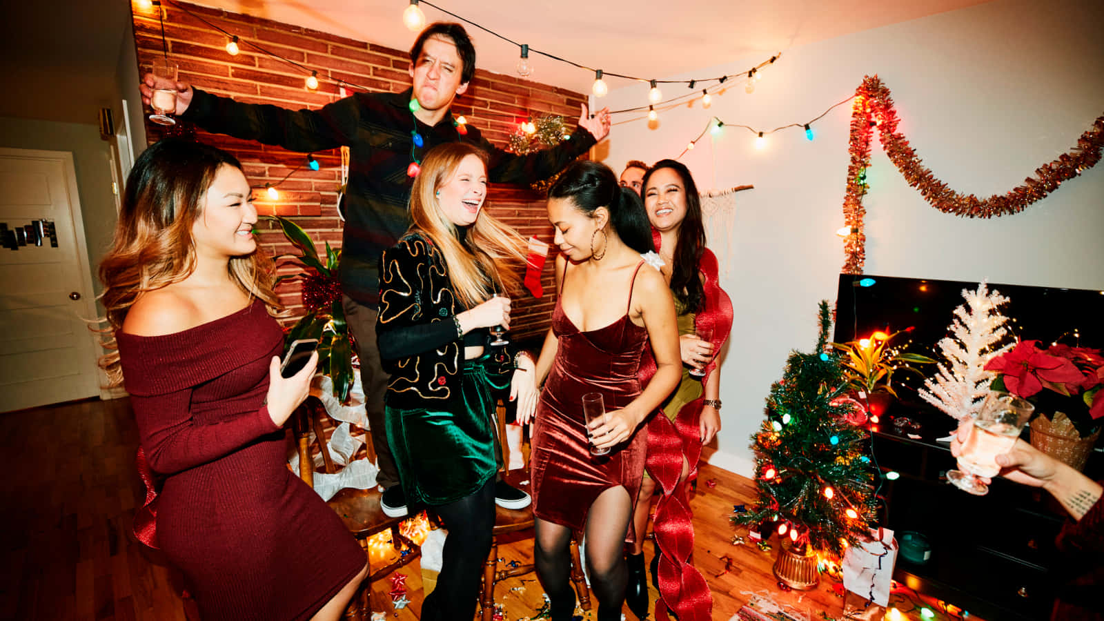 女生聖誕派對照片 壁紙 — 朋友們在舒適的聖誕派對上歡笑並舉杯暢飲，派對由燈串和裝飾好的聖誕樹照亮。