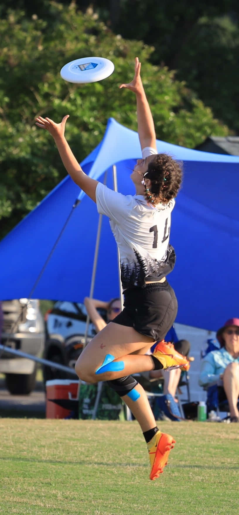 女運動員接飛盤 Android Ultimate Frisbee 背景 壁紙 — 一名女運動員在空中張開雙臂跳起，接住飛行的飛盤，背景是綠樹和藍色帳篷。