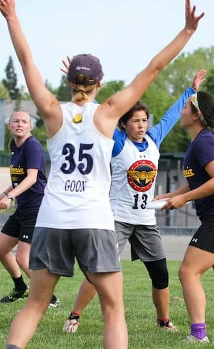 女運動員防守姿勢舉臂Android Ultimate Frisbee背景 壁紙 — 一位身穿白色球衣、背後印有35號和'GOON'字樣的女飛盤選手在草地上舉臂慶祝勝利。