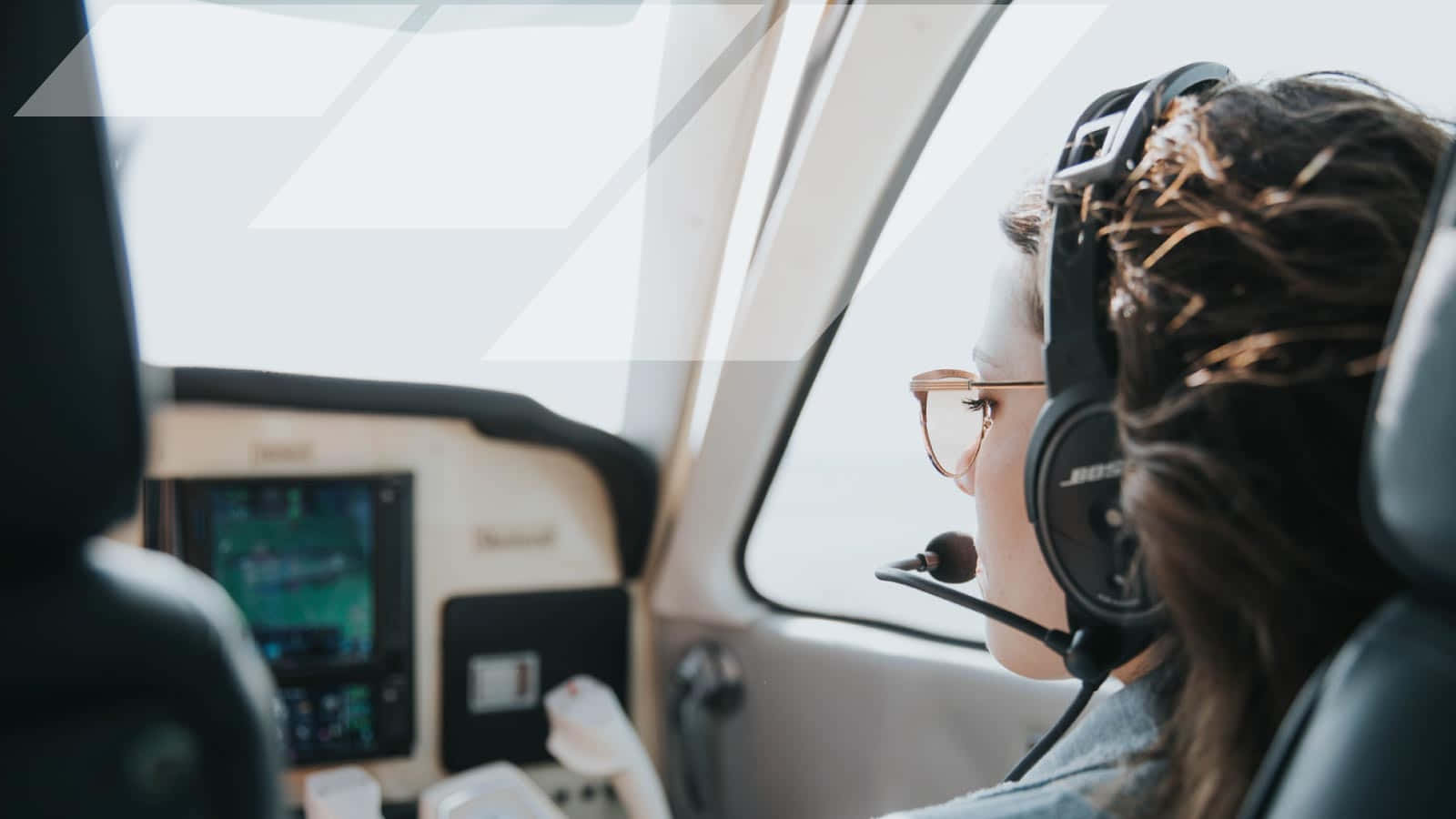 女駕駛員導航飛行 壁紙 — 一名戴著耳機和眼鏡的機師專注地坐在小型飛機的駕駛艙內，望向窗外。