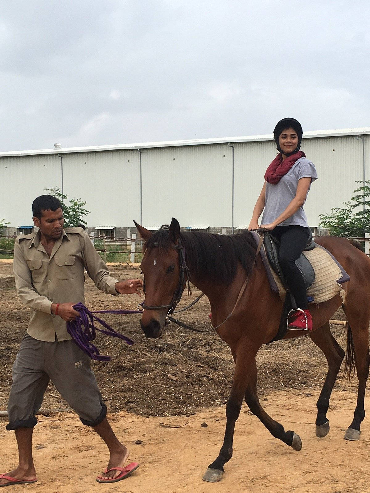 女騎手悠閒策馬，助手牽引馬匹 壁紙 — 一名女子微笑騎著一匹棕色馬，由一名男子握著韁繩在白色建築附近的塵土小徑上引導。