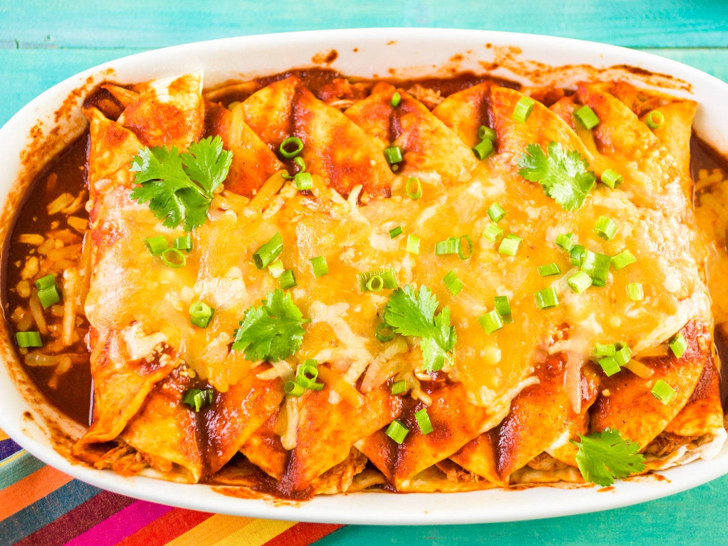 奶酪與香料 enchiladas 盤 壁紙 — 金黃色的玉米捲餅浸在紅醬、融化芝士和新鮮芫荽中，放在白色碟子上，置於色彩繽紛的條紋布上。