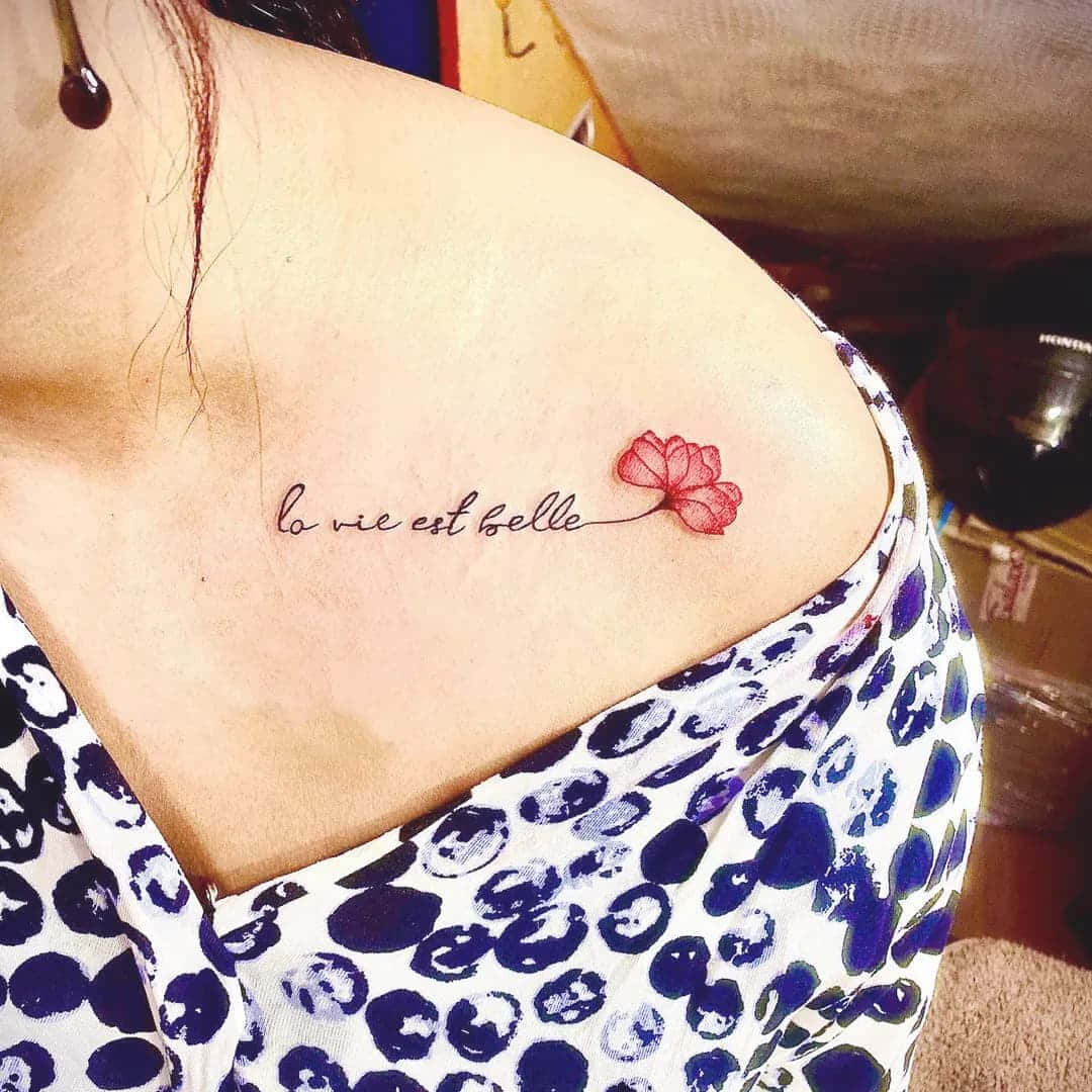 她有風格 壁紙 — 一個精緻的紅色花朵紋身，帶有草書法文文字 la vie est belle，裝飾在一個人穿著圖案上衣的肩膀上。