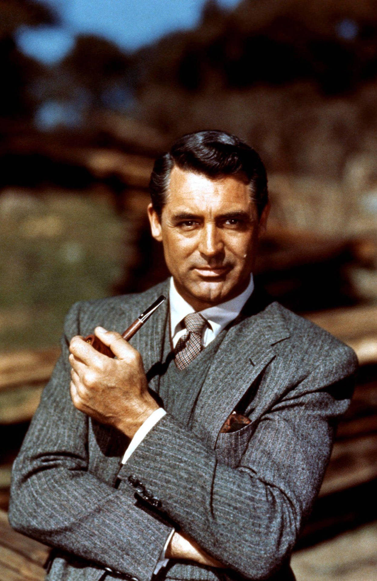 Cary Grant 身穿灰色西裝，在戶外陽光下手持煙斗。